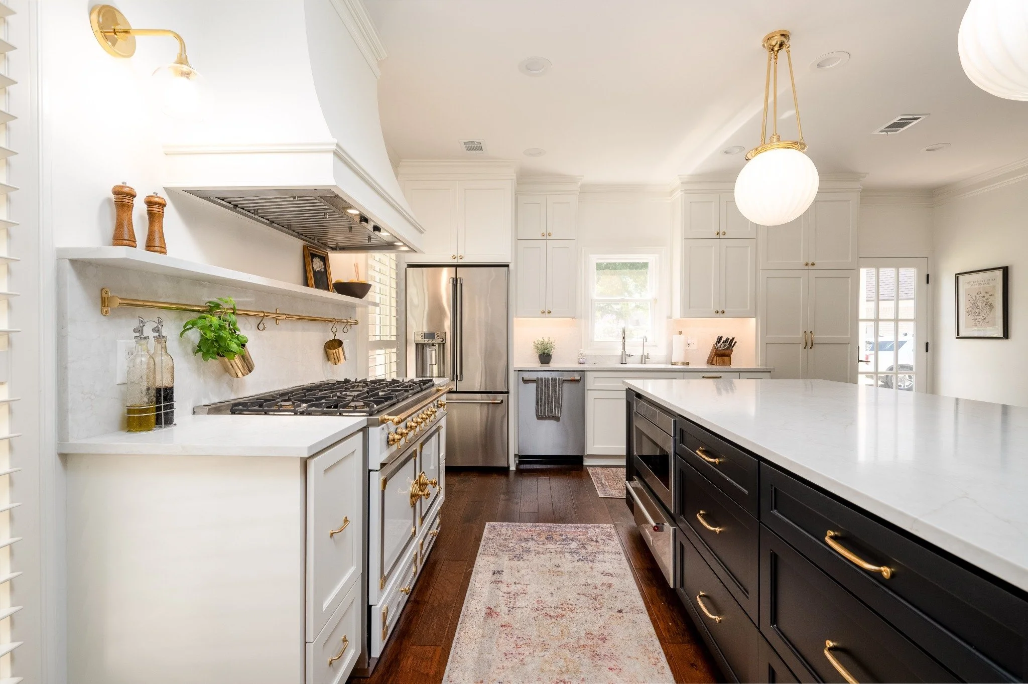 Modern kitchen with white and black cabinetry, stainless steel appliances, and a central island with a white countertop. There are pendant lights hanging from the ceiling, and a window above the sink lets in natural light. The kitchen has dark wood flooring and decorative items on the shelves.