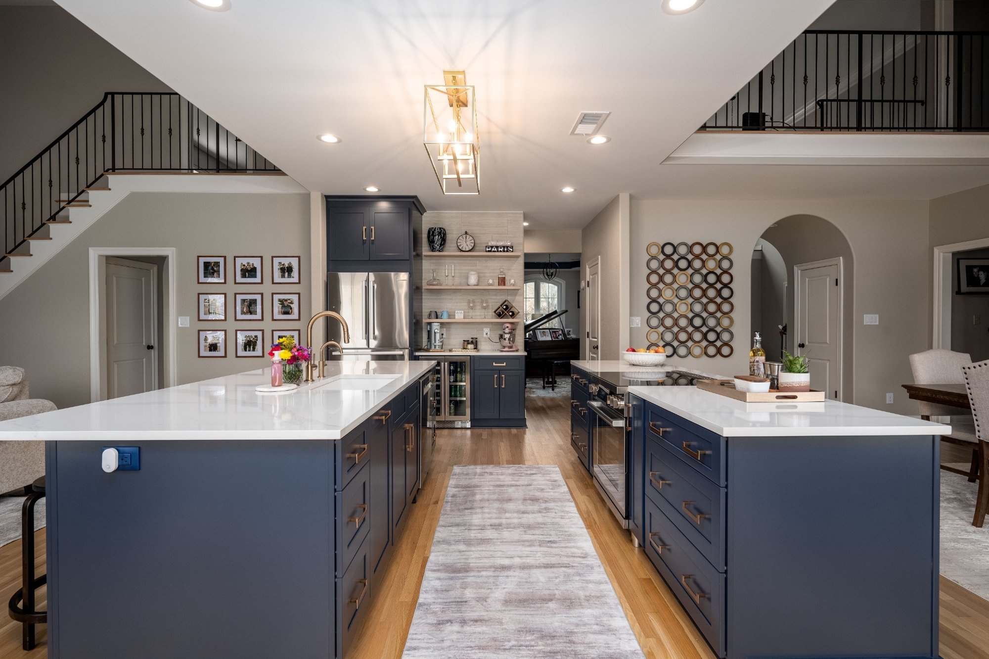 Modern kitchen with blue cabinets, white countertops, and wooden flooring. Features include a large island, a wine cooler, wall shelves with decor, and a light fixture on the ceiling. Part of a living area with framed photos and a staircase railing visible.