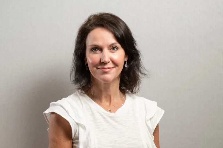 A woman with shoulder-length dark hair and a white top, standing against a plain gray wall.