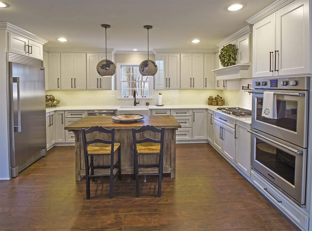 Bright modern kitchen with white cabinets, stainless steel appliances, and a wooden island with two black chairs. Recessed lighting and pendant lights hanging above the island. Window over the sink with a tree visible outside. Decor includes potted plant, baskets, and a large tray on the island.