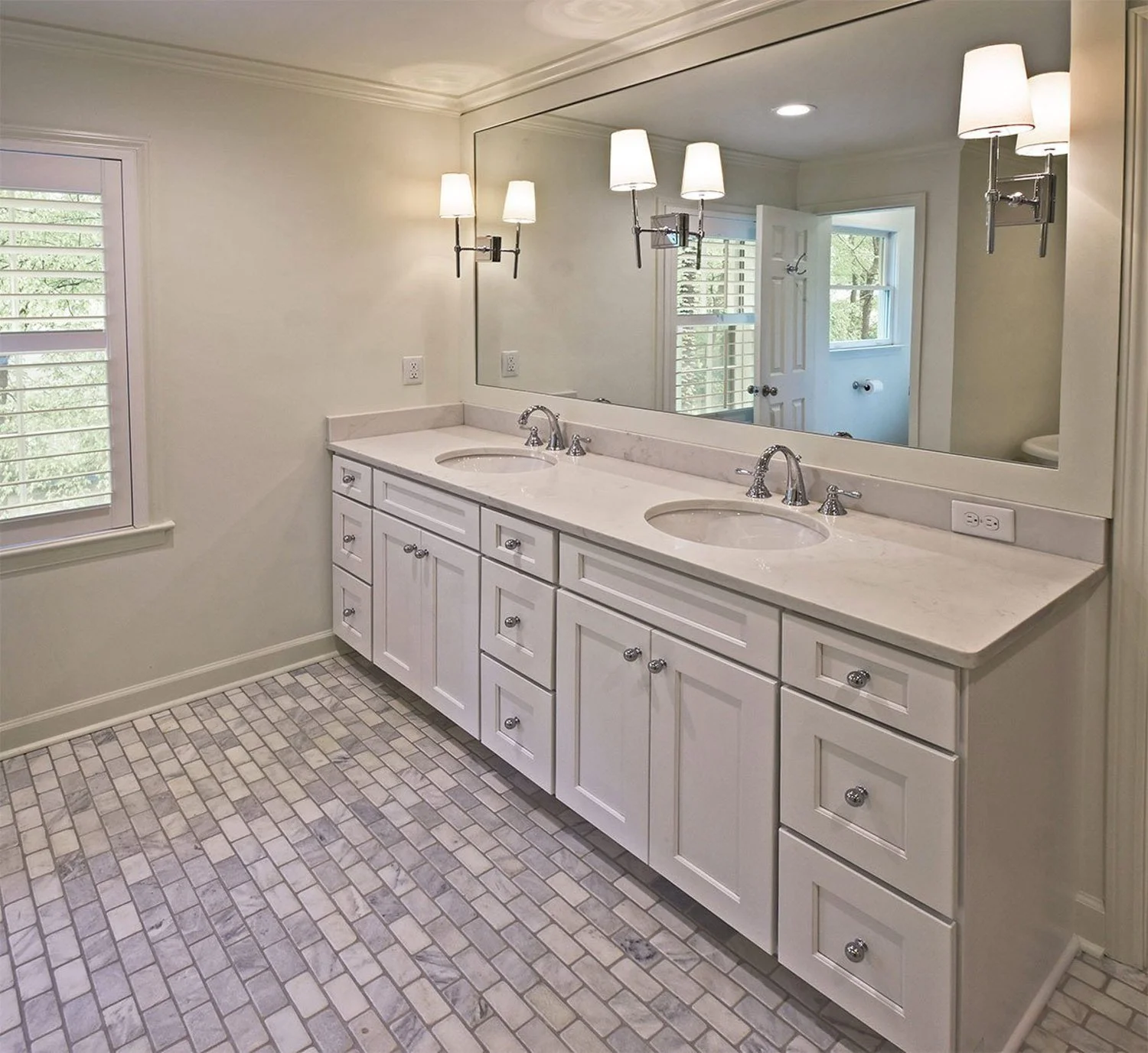 The custom double vanity includes white shaker cabinetry, marble countertops, polished chrome hardware, and decorative lighting that brightens the entire space.