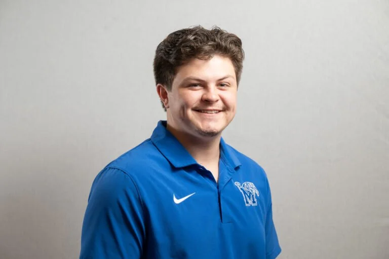 Portrait of a young man smiling, wearing a blue Nike polo shirt with a logo that has a large letter 'M' and a smaller, intertwined 'F' on the chest, standing against a plain light gray background.