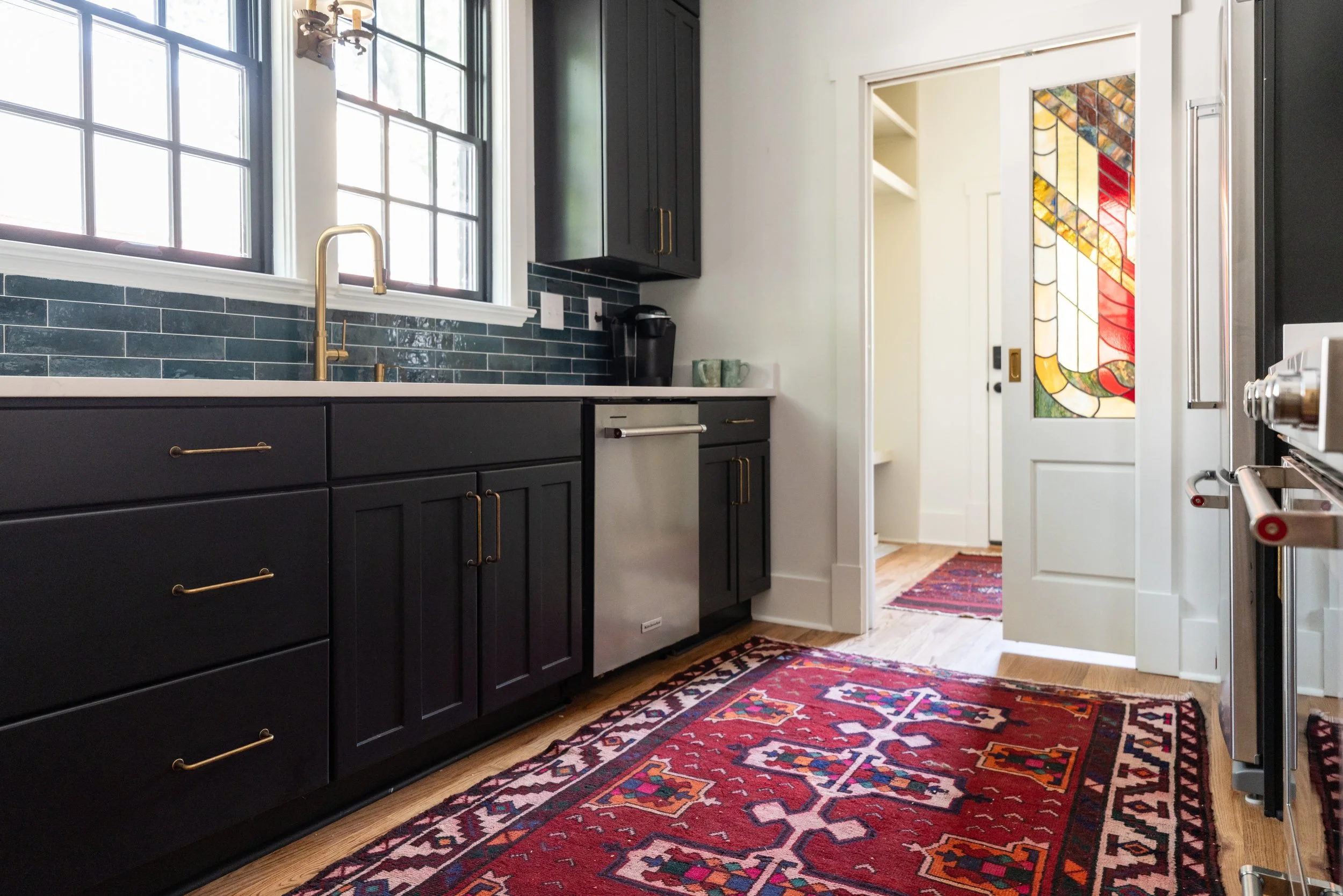 A modern kitchen with black cabinets, gold handles, large windows, a colorful stained glass door, and a patterned red runner on a wooden floor.