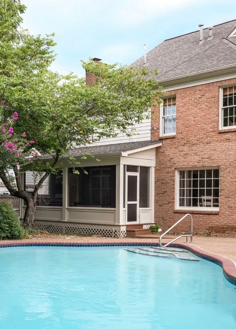 Backyard view with a swimming pool, a brick house with white-framed windows, a screened porch, and a tree with purple flowers.
