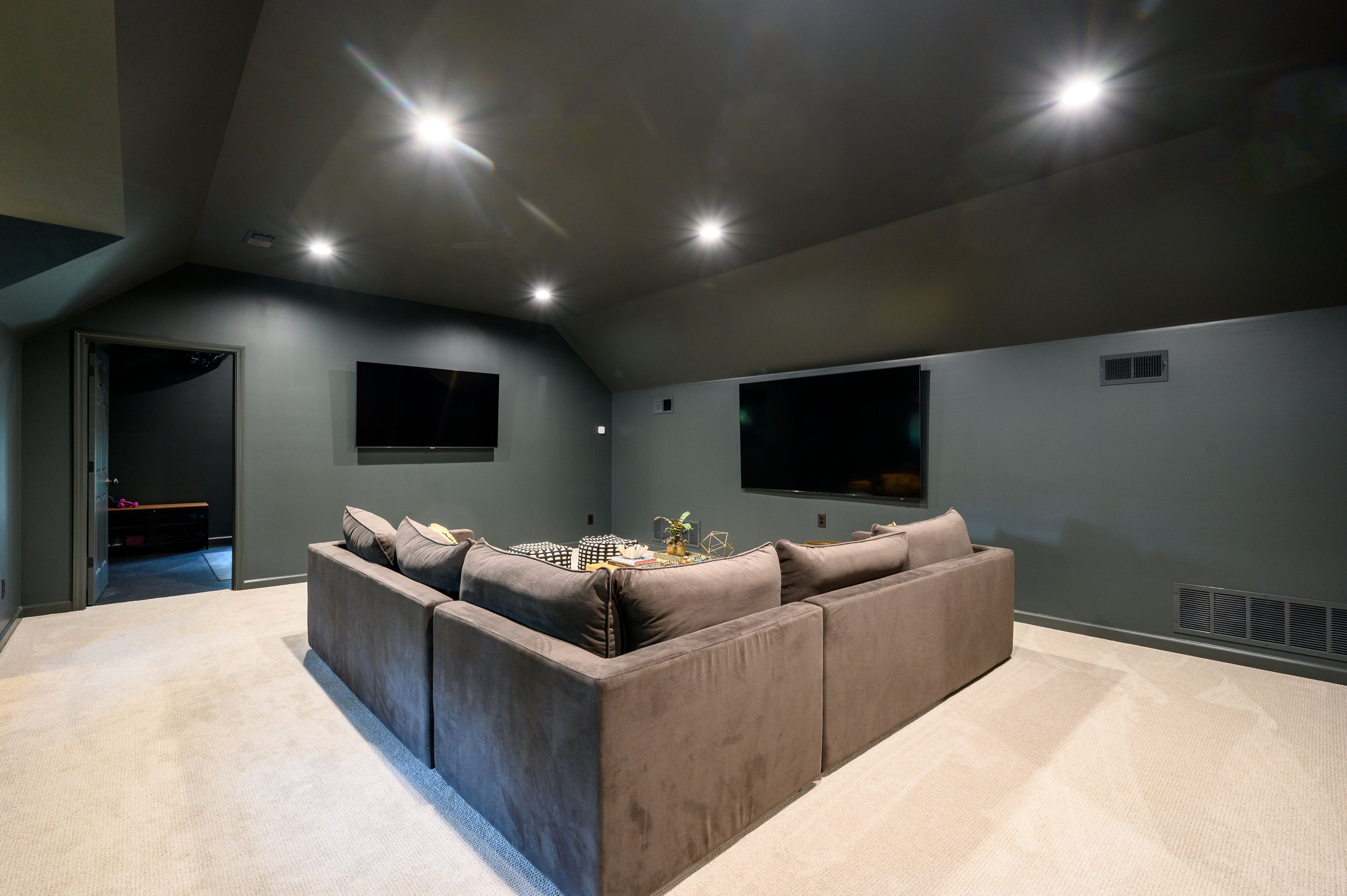 A cozy living room with a large L-shaped gray sofa, two mounted flat-screen TVs on a gray wall, and recessed lights on the ceiling.