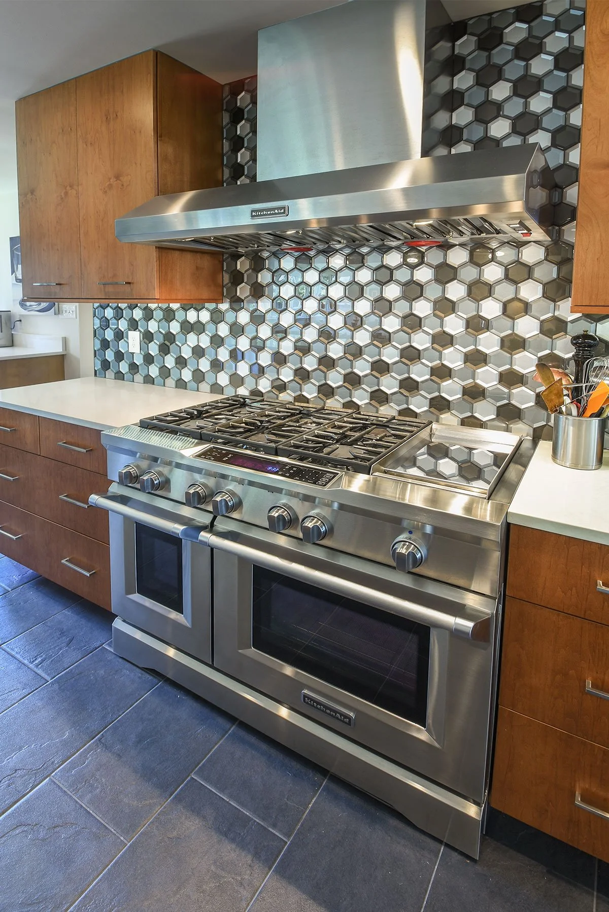 Modern kitchen stove with stainless steel oven and burners, wooden cabinets, hexagonal tile backsplash, and a metal range hood.