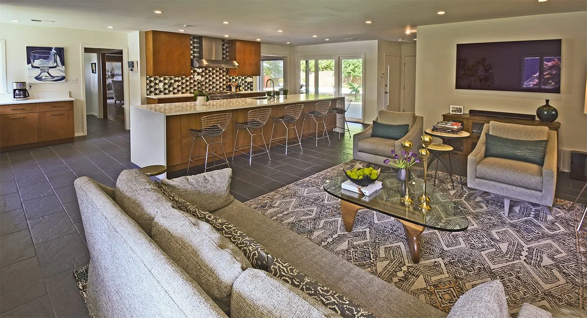 Living room with a kitchen island, bar stools, a TV, two armchairs, a sofa, a glass coffee table, and decorative accents.