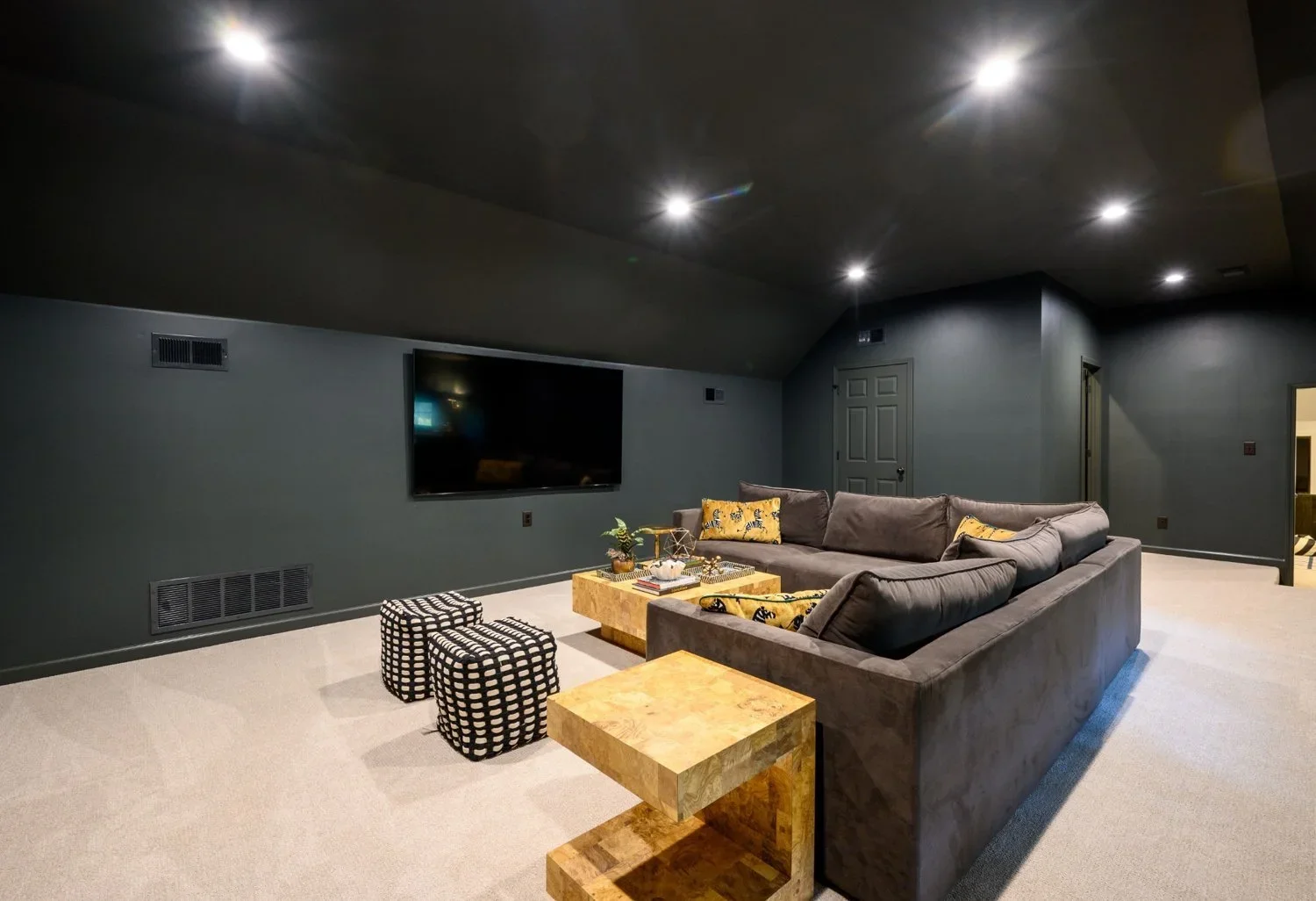 Living room with dark green walls, a large gray sectional sofa with yellow accent pillows, a black TV mounted on the wall, small black and white checkered ottomans, and a wooden coffee table, in a room with a ceiling-mounted light and beige carpet.