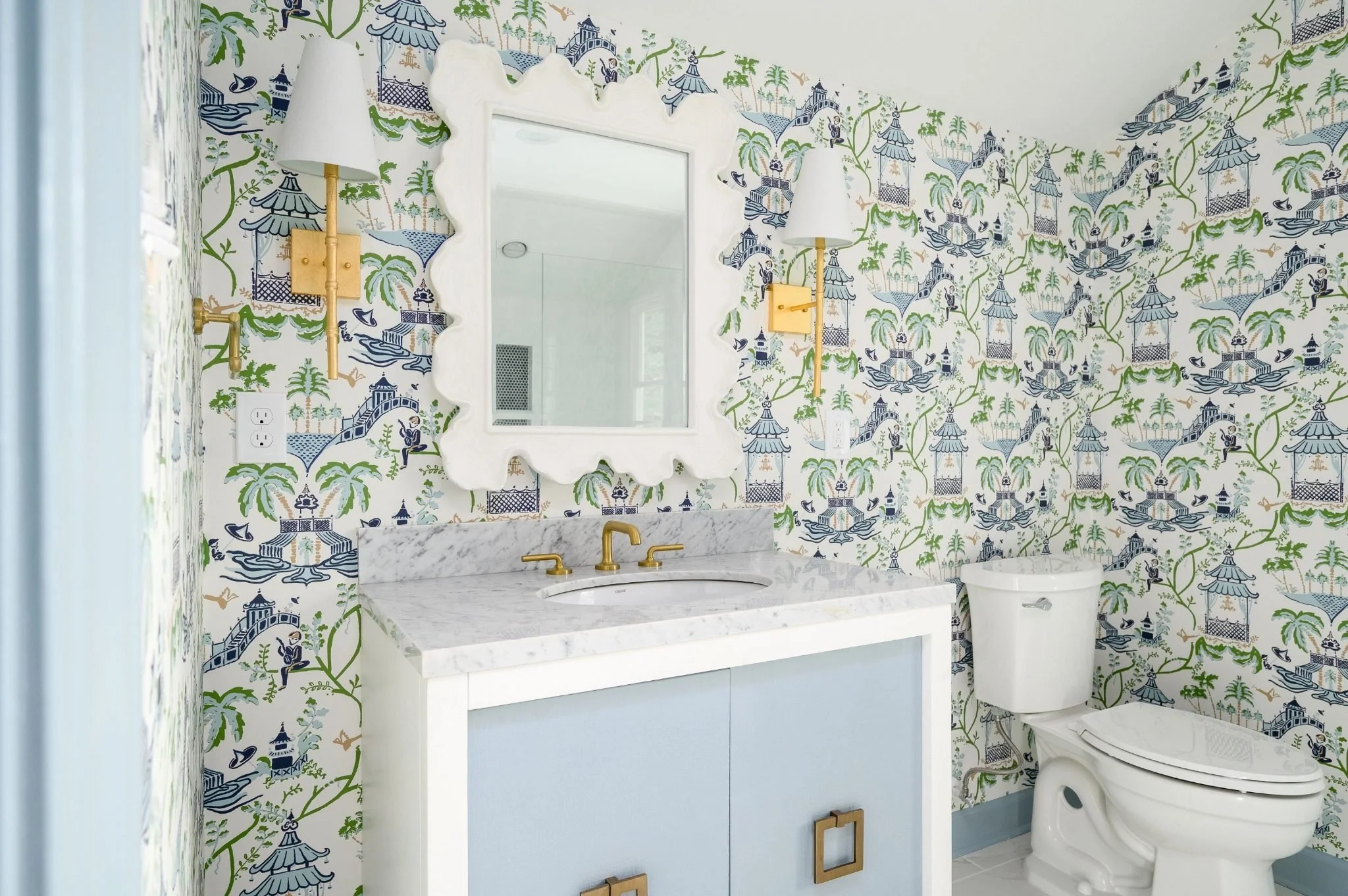 Custom powder room featuring blue vanity cabinetry, a white marble countertop, brass faucet and sconces, a decorative scalloped mirror, and blue-and-green patterned wallpaper for a bold, designer-inspired look.