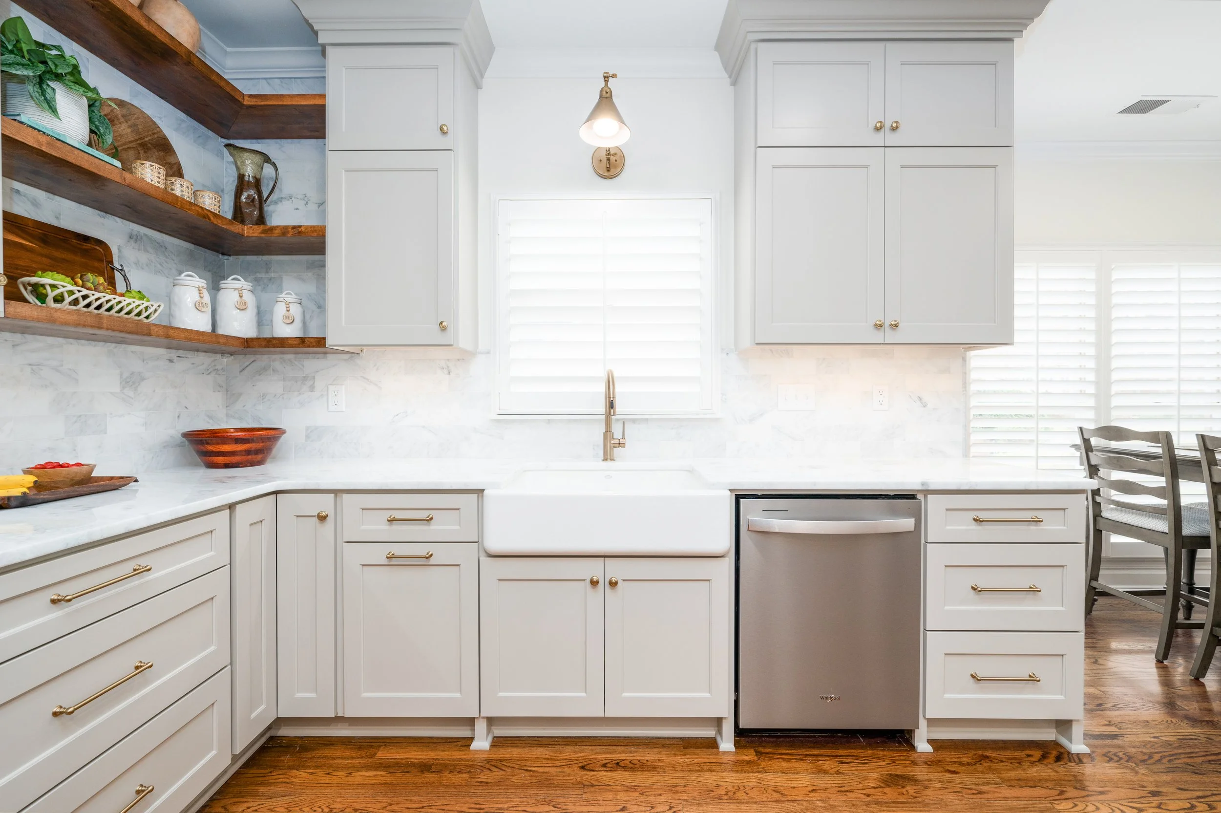 A bright kitchen with white cabinetry, marble countertops, open wooden shelves with decorative jars, a gold faucet, and large windows with white shutters.