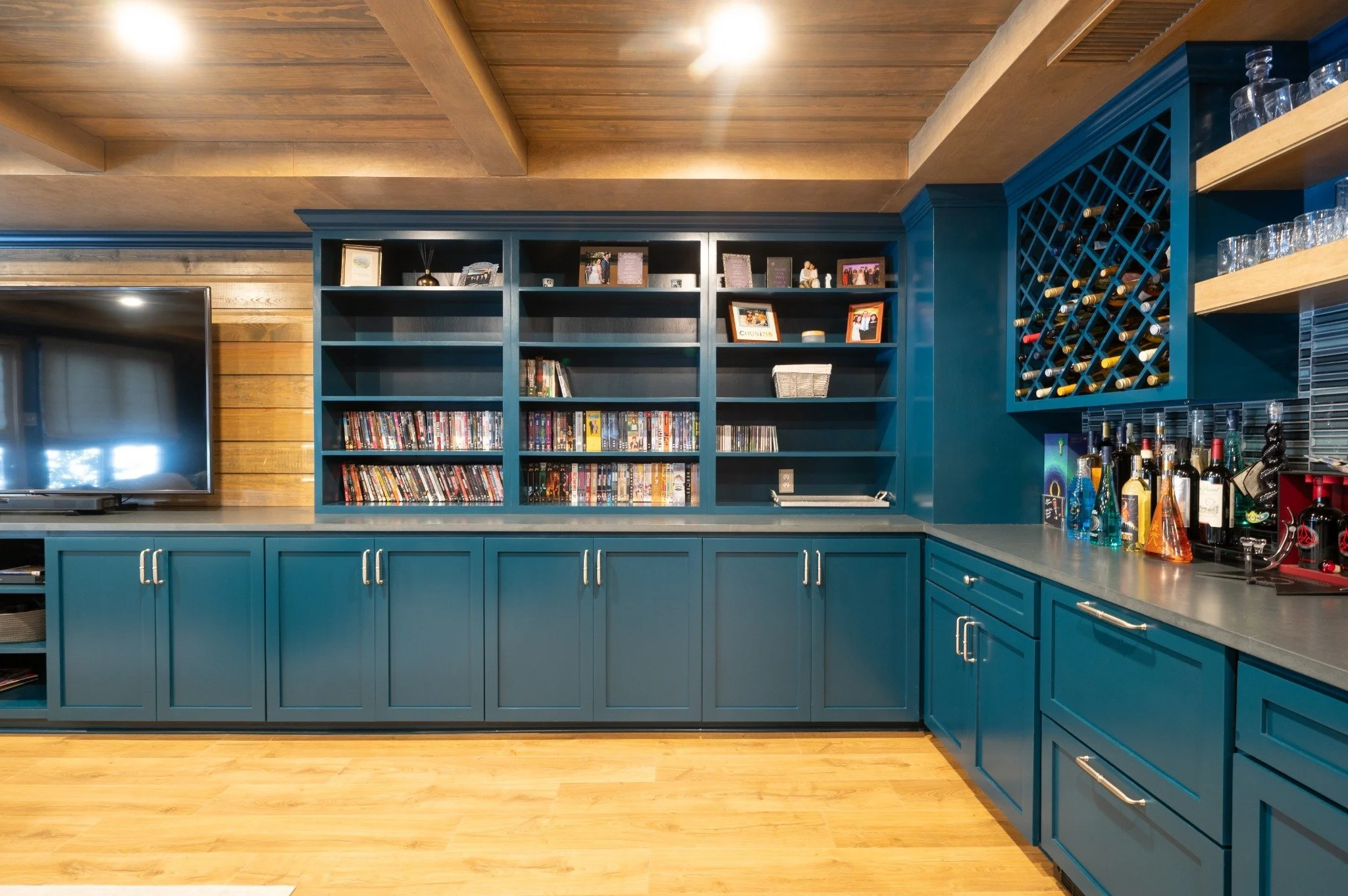 Interior of a kitchen with blue cabinets and open shelving. The upper shelves display framed photos and books, while the counter holds various bottles and barware. The wall behind the shelves is wood-paneled, and there is a flat-screen TV on the left side of the image.