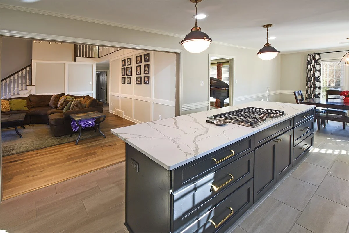 Open-concept kitchen and living room with a large kitchen island with a white marble countertop, dark cabinetry with gold handles, and two pendant lights hanging above. In the background, there is a sitting area with a brown sectional sofa, a staircase, and a wall decorated with framed photos. To the right, a dining table with chairs and a window with patterned curtains can be seen.