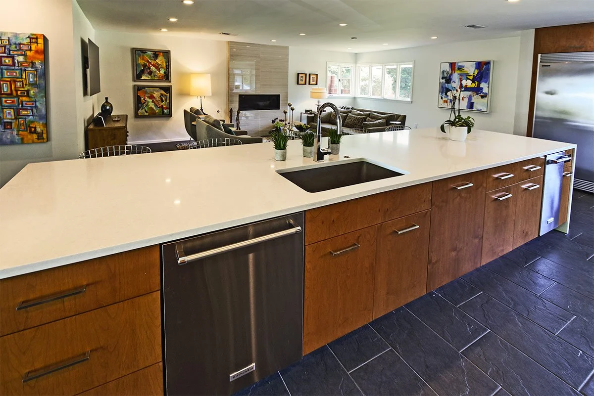 Modern kitchen island with white countertop, built-in sink, and potted plants, overlooking a living room with abstract artwork and large windows.