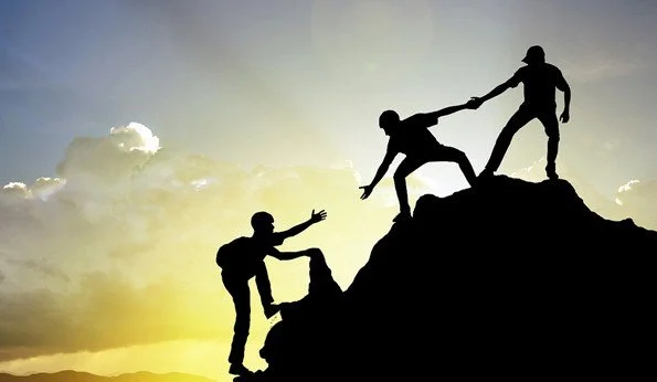 Silhouettes of two people helping each other climb a mountain against a sunset sky.