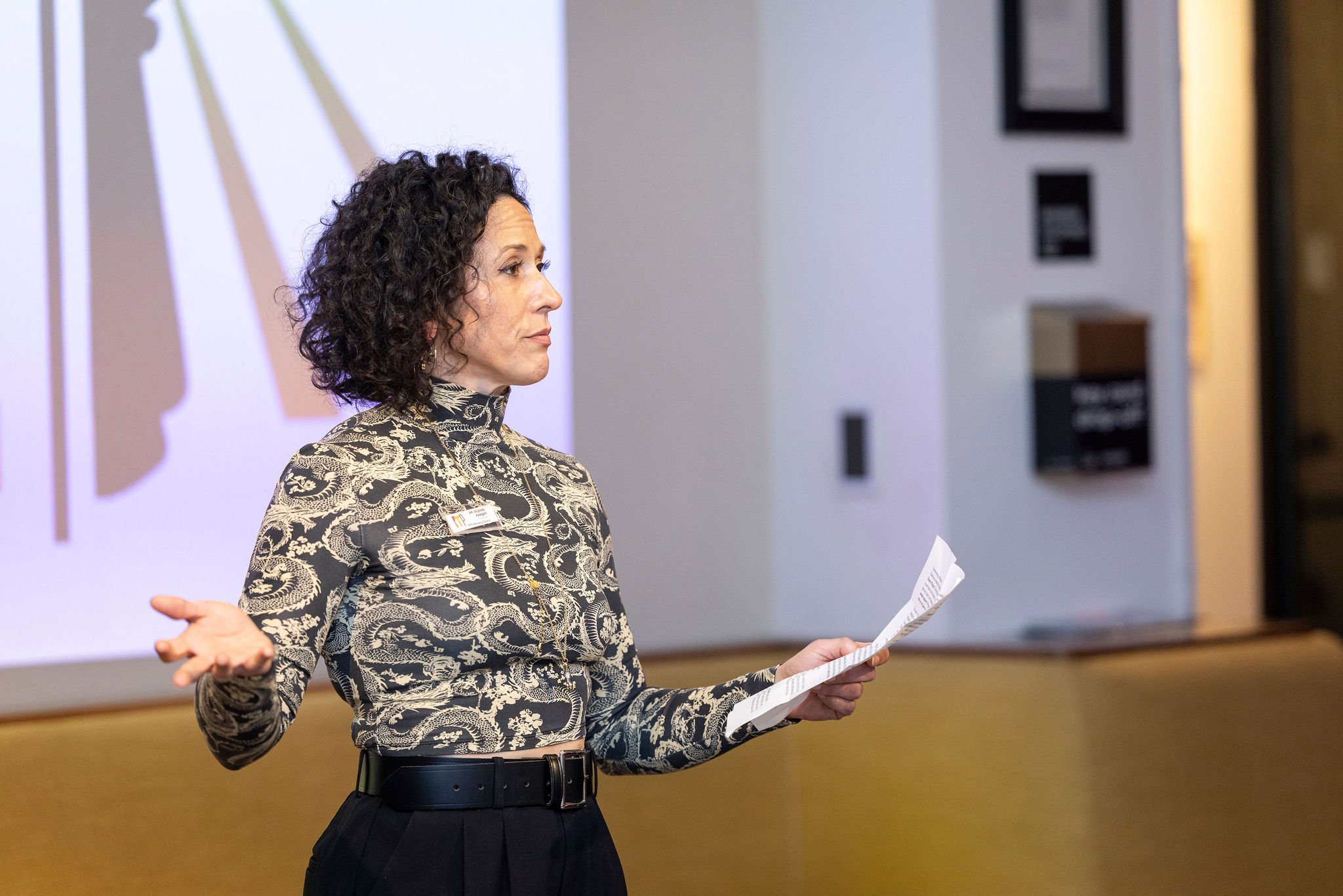 A woman with curly dark hair, wearing a patterned blouse and black pants, stands in front of a presentation screen, holding papers in her right hand, gesturing with her left hand.