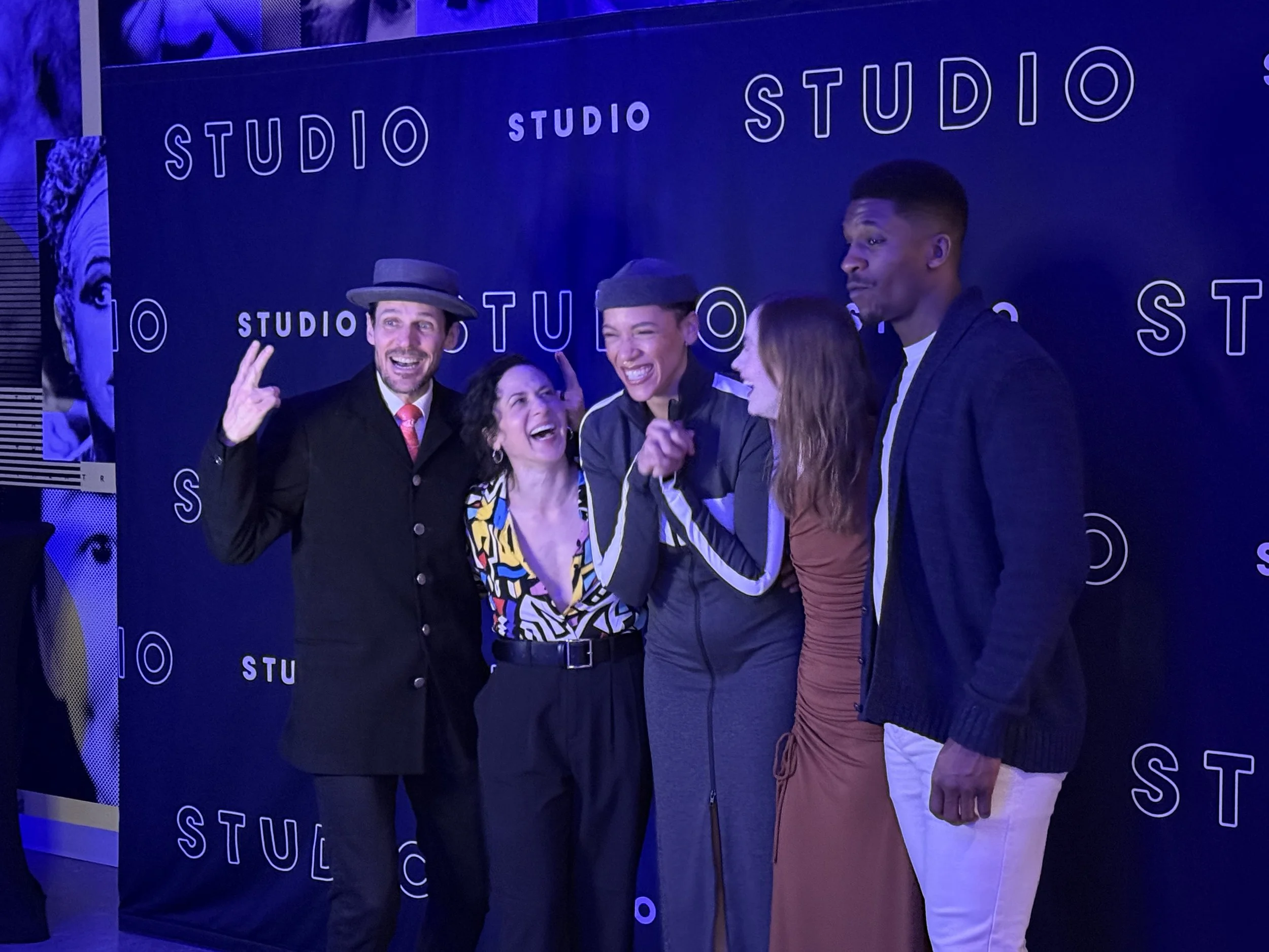 Five people posing in front of a paper backdrop that reads 'STUDIO' multiple times, smiling and laughing. The group includes two women and three men, dressed in casual and stylish clothing.