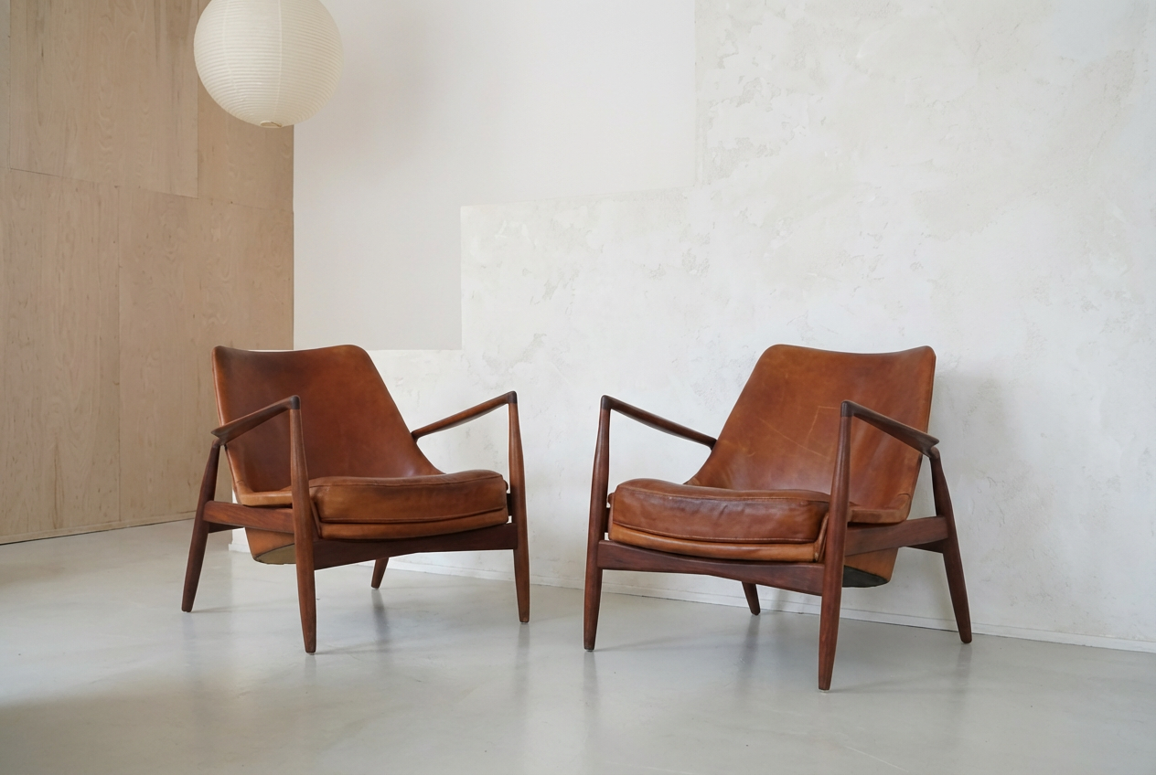 Two vintage leather armchairs with wooden frames in a minimalist room with white walls and a paper lantern hanging from the ceiling.