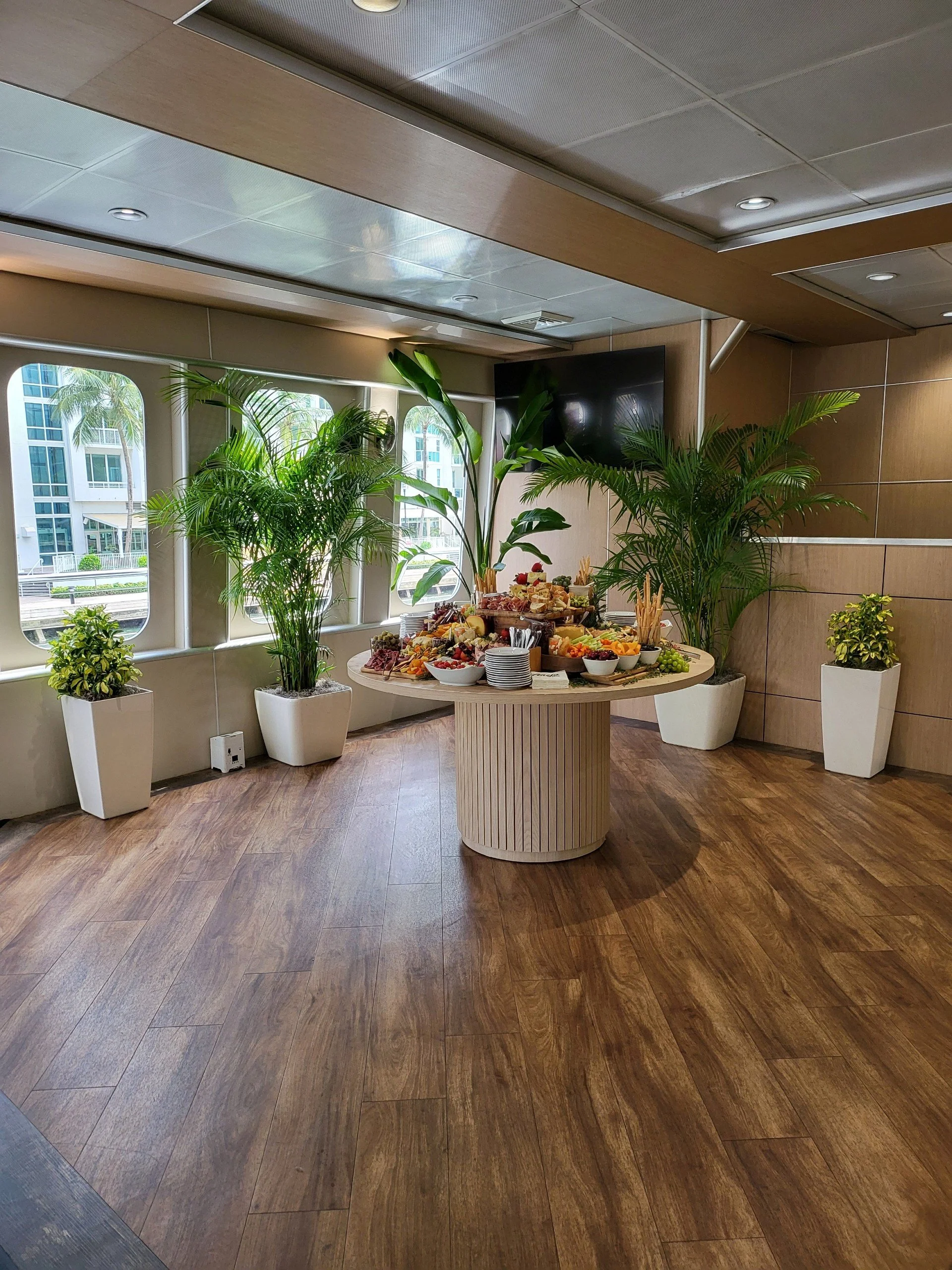 A buffet table with various food items and fruits, surrounded by potted plants, in a well-lit room with wood-flooring and large windows.