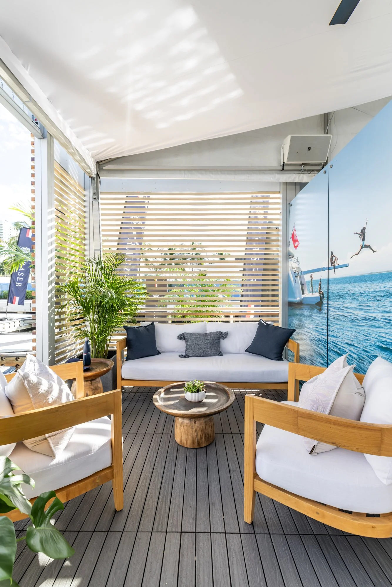 Outdoor seating area with white cushioned wooden furniture, black and gray pillows, a small round wooden coffee table with a potted plant, green plants, and a large mural of a person diving off a boat into water with a clear sky.