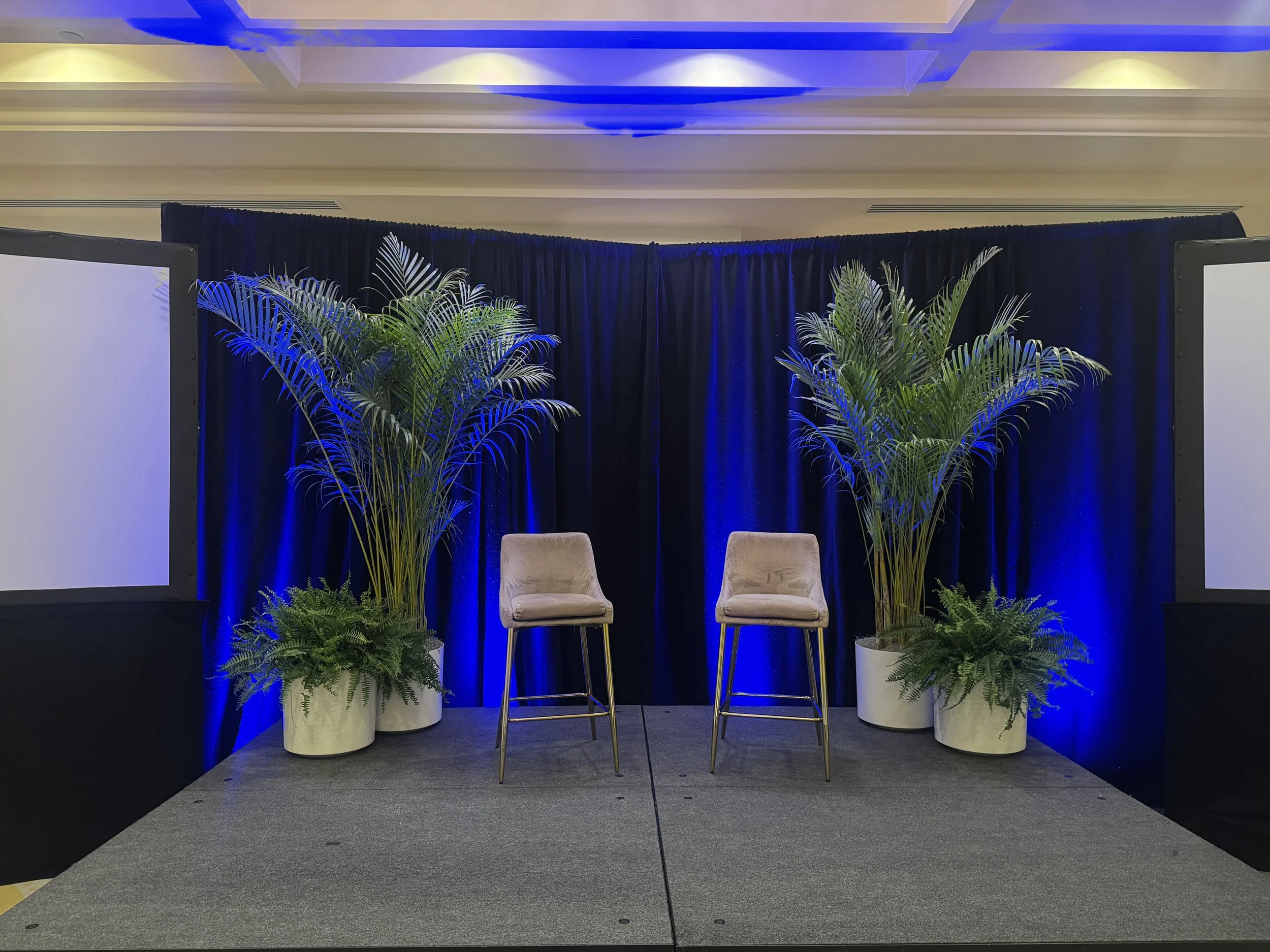 Stage setup with two beige chairs, potted plants, dark blue curtains, and two large screens on either side.