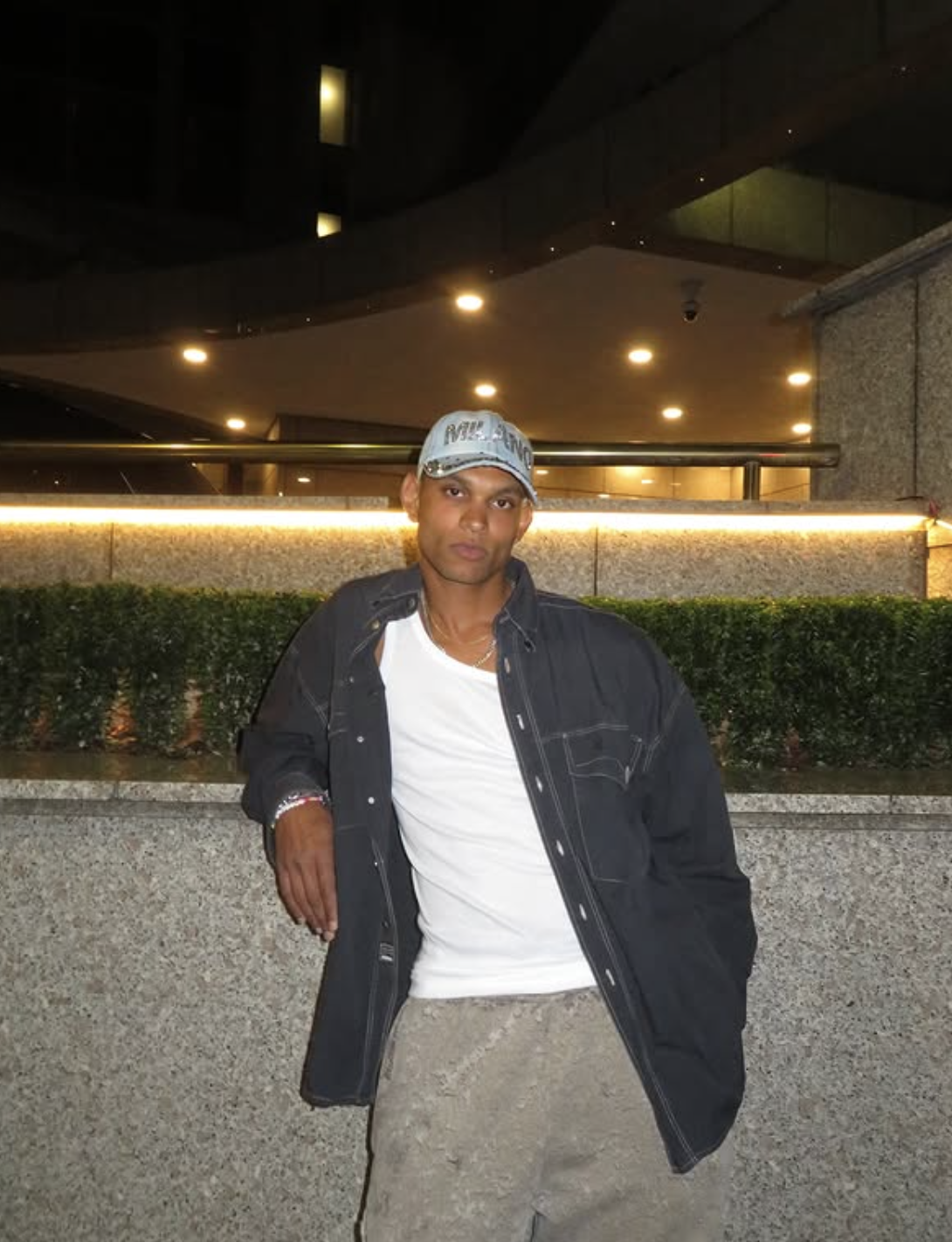 A young man standing outdoors at night, wearing a baseball cap, white T-shirt, black jacket, and beige pants, leaning against a concrete ledge with greenery behind him, illuminated by city lights.