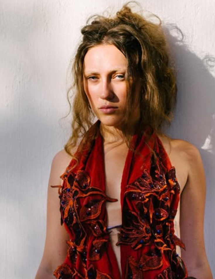 Woman with curly, frizzy hair wearing a red, decorative scarf or shawl against a plain background.