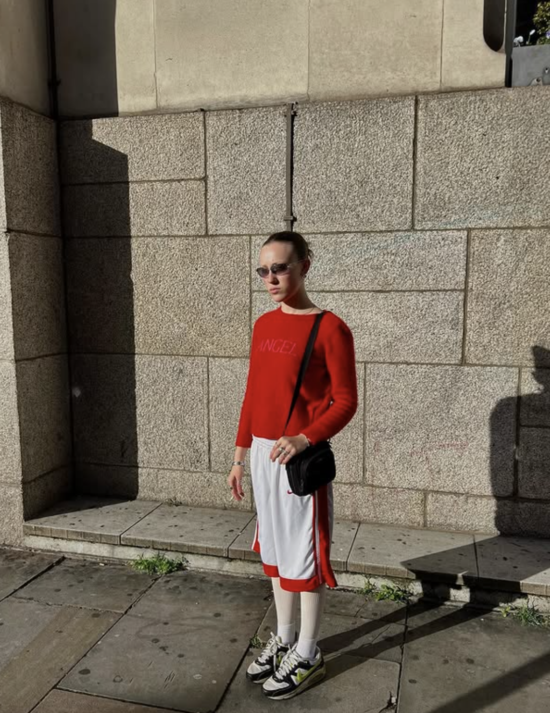 A woman with short dark hair wearing sunglasses, a red sweater, white shorts with red stripes, white tights, and sneakers, carrying a black crossbody bag, standing on a sidewalk against a beige stone wall in sunlight.