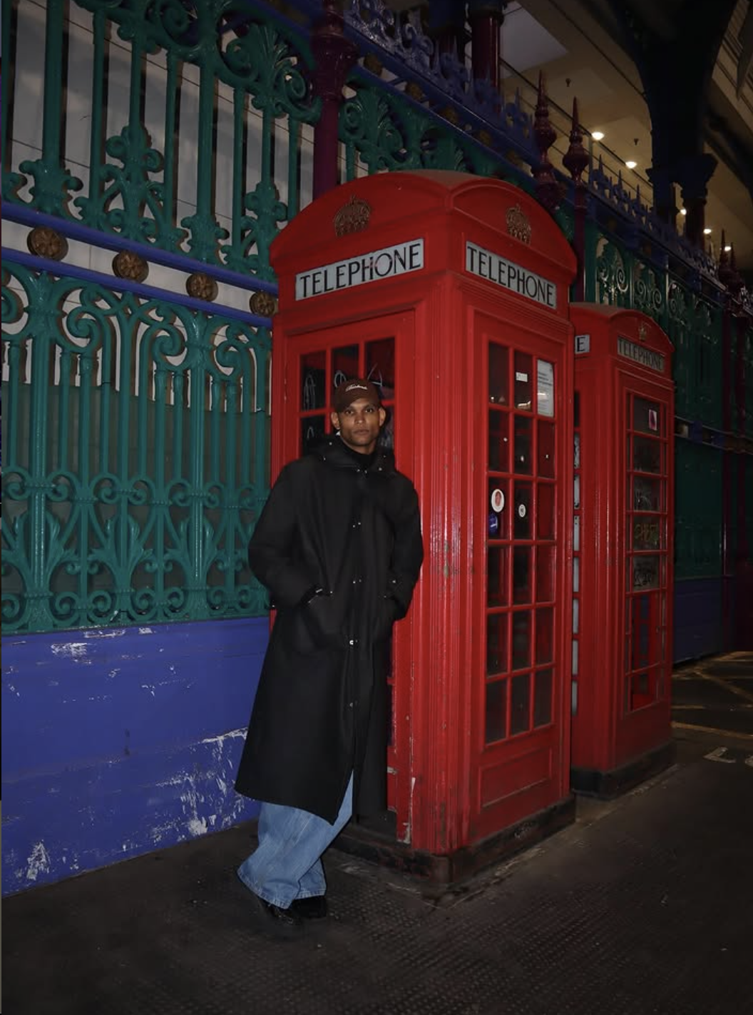 A person in a black coat and blue jeans standing next to red telephone booth in front of a decorative wrought iron fence at night.