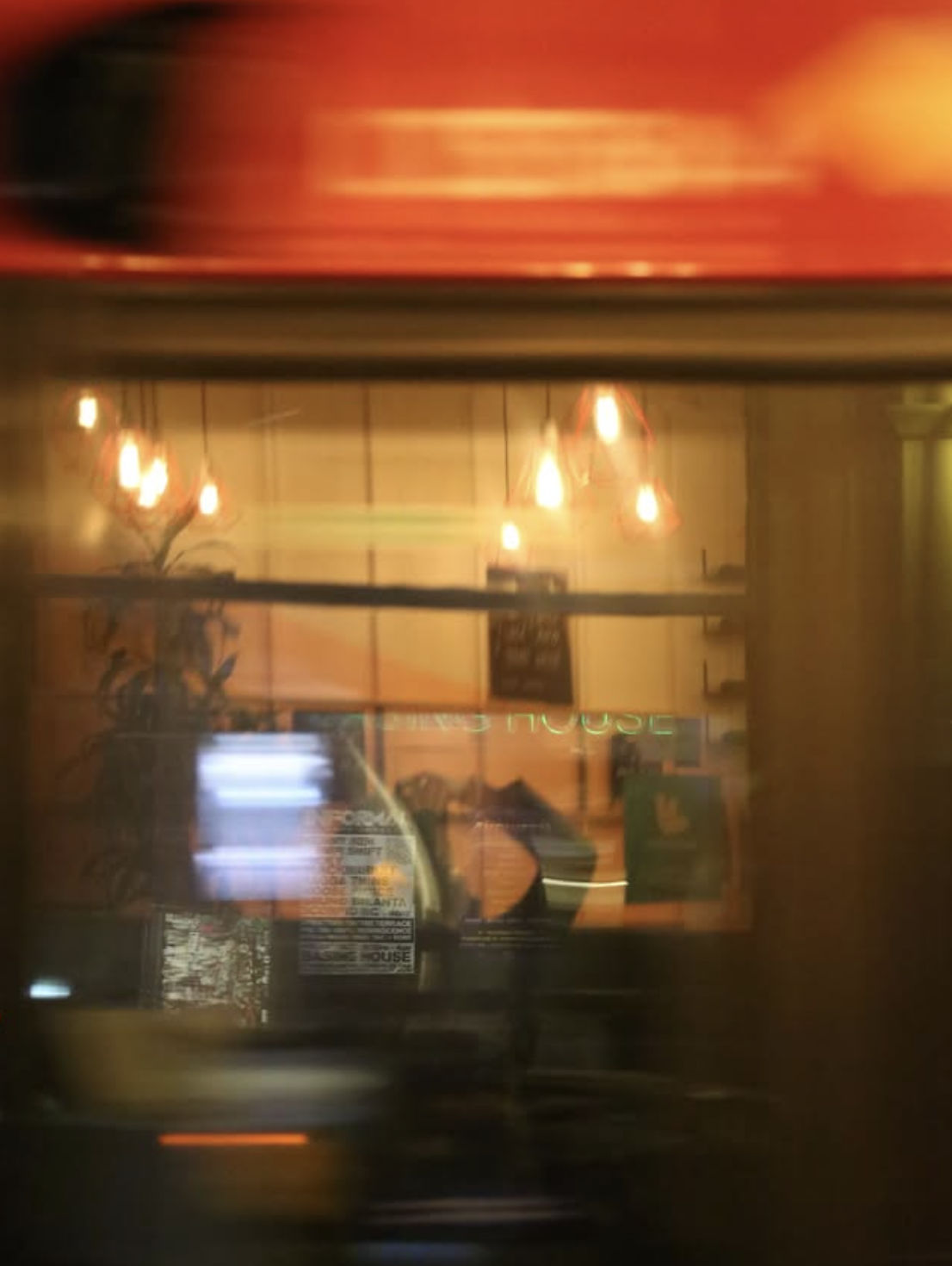 A blurry view of a restaurant or cafe interior through a window, with hanging pendant lights and a person taking a selfie.
