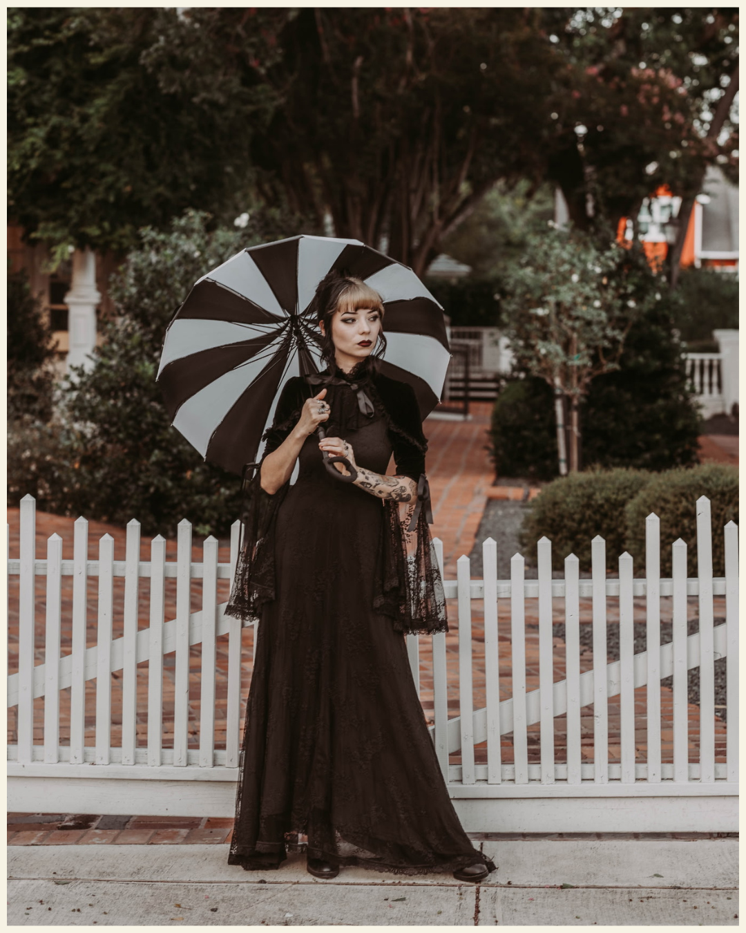 A woman dressed in black gothic fashion holding a black and white umbrella, standing in front of a white picket fence with a garden and brick pathway in the background.