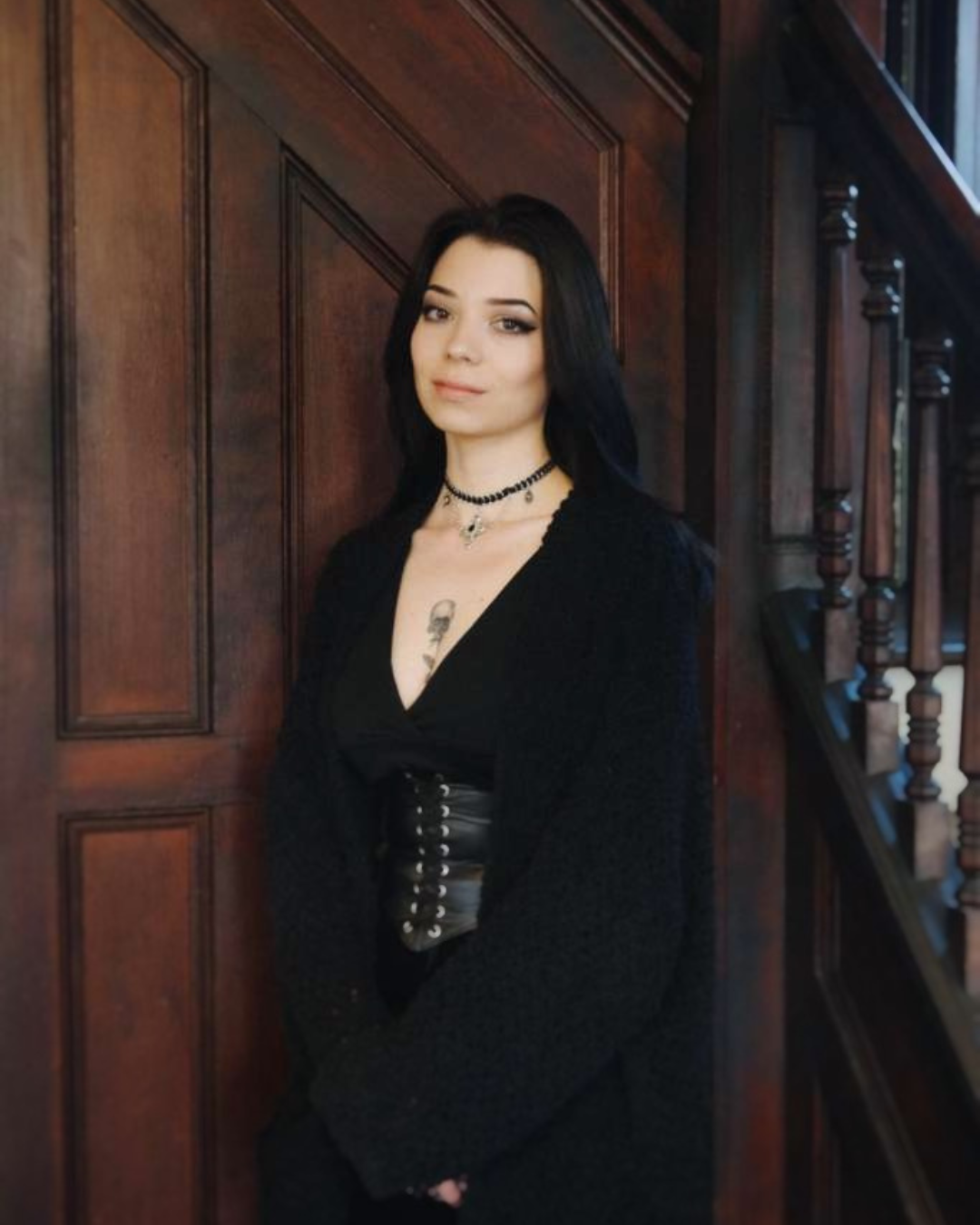 A young woman with black hair and fair skin stands indoors against a wooden wall, wearing a black V-neck top, a black jacket, a black corset, layered necklaces, and has a tattoo on her chest.