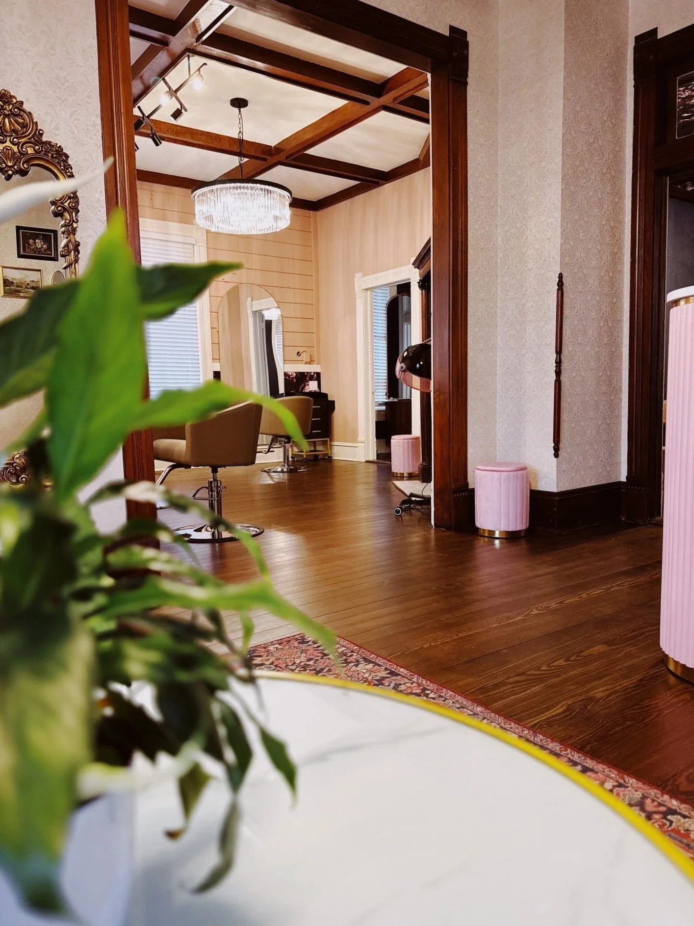 Interior view of a room with wooden floors, a doorway, pink ottomans, and a vanity area with a chair and mirror. There are vintage-style lamps and a chandelier visible.