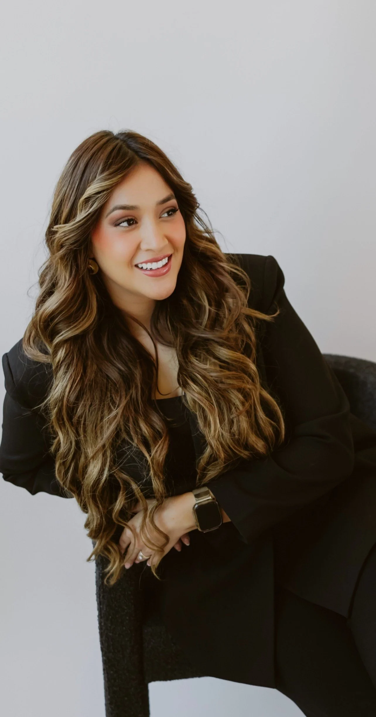 Young woman with long, wavy brown hair and a bright smile, sitting on a black chair against a plain white background, wearing a black blazer and a smartwatch.