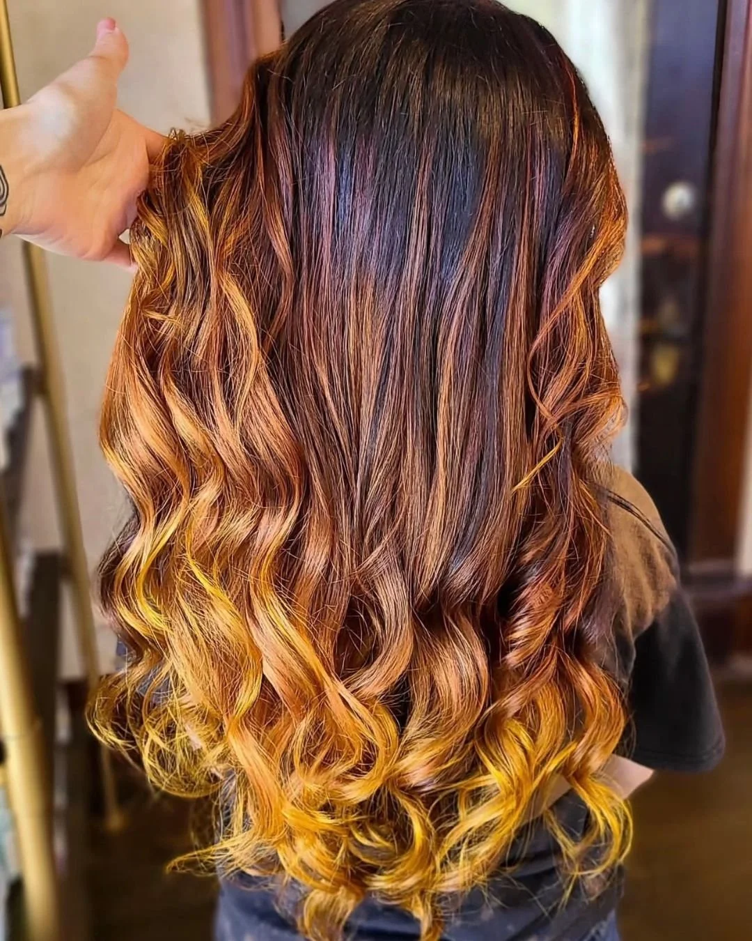 Back view of a woman with long, curly hair that transitions from dark brown at the roots to vibrant orange and yellow at the tips.