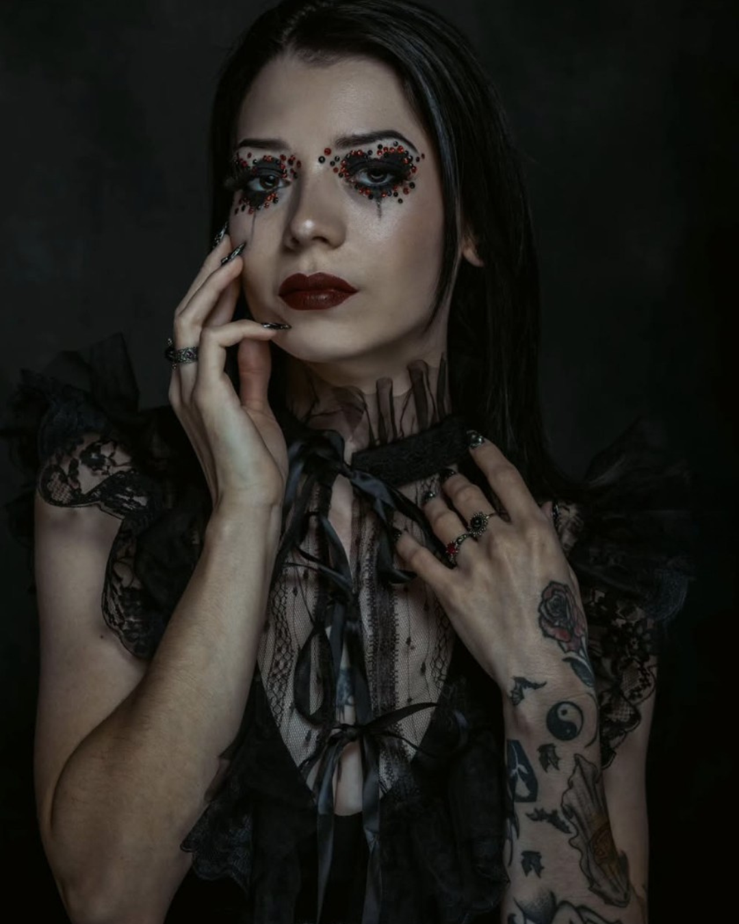 A woman with dark makeup, black lipstick, and dramatic eye makeup decorated with red and black rhinestones, wearing black lace clothing, and tattoos on her arm, set against a dark background.