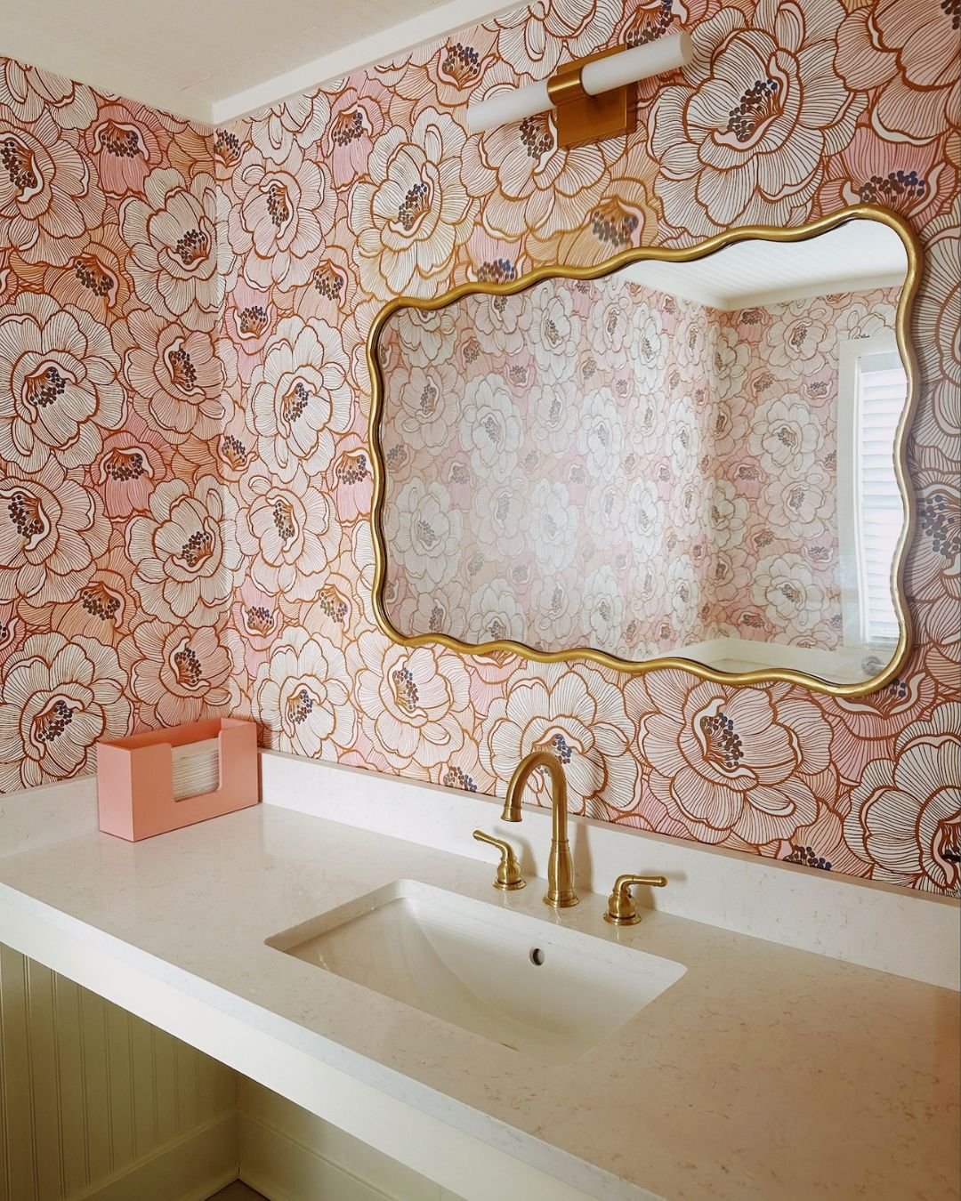 A bathroom with a pink floral wallpaper, a white countertop with a sink, gold faucet, and a decorative mirror. A pink box and a soap dispenser are on the counter. There is a trash bin and tiled floor.