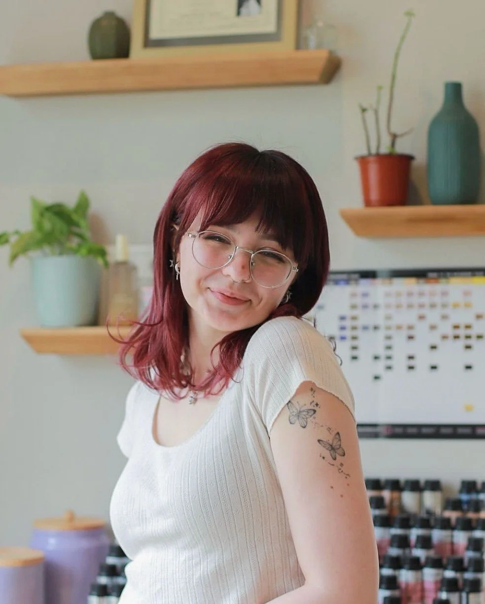 A young woman with red hair and glasses smiling in a room with shelves and potted plants in the background.