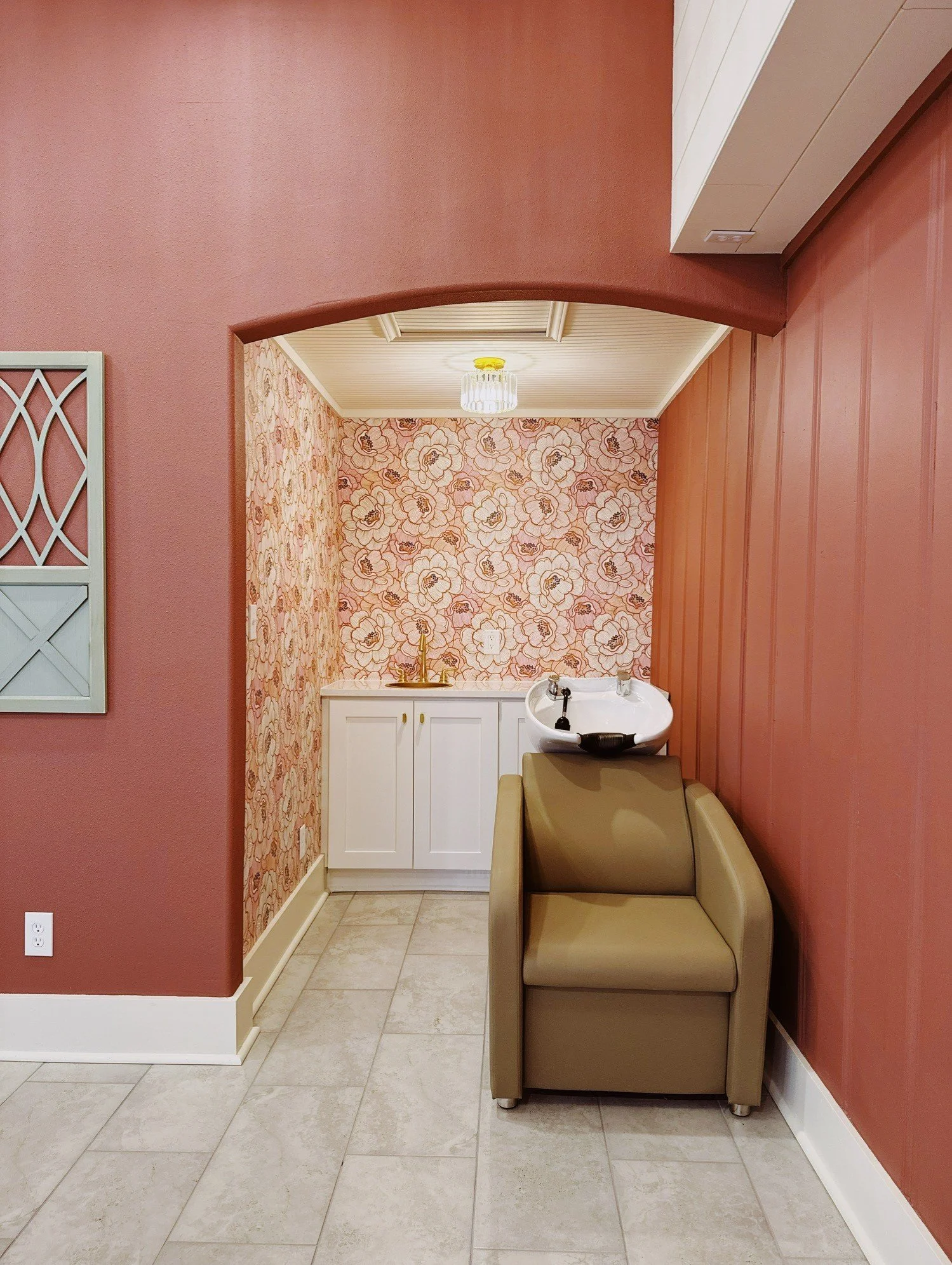 Interior of a room with a pink wall, a small sink with gold fixtures, floral wallpaper, a beige salon chair with a white hair wash basin, white cabinets, and tiled floor.