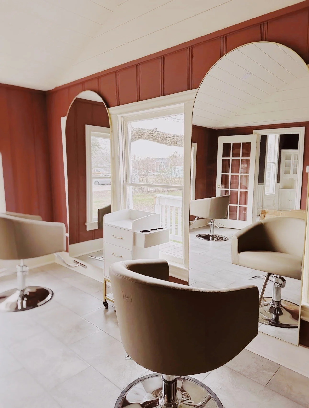 Empty hair salon with beige salon chairs, mirrors, and large windows in a room with wooden wall paneling.