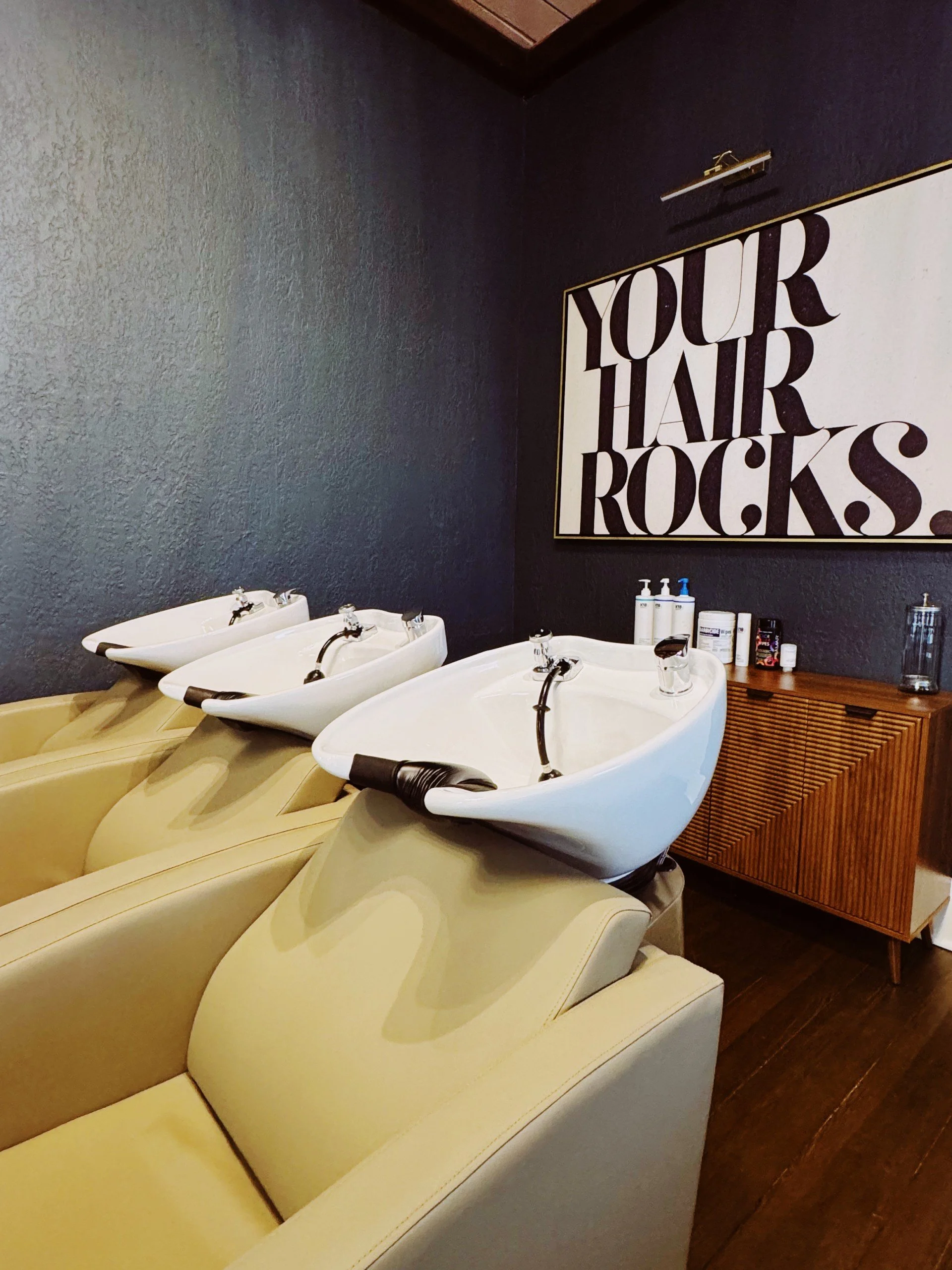 Three salon wash basins with yellow chairs underneath and a wooden cabinet in the background, with a sign reading 'Your Hair Rocks' on the wall.