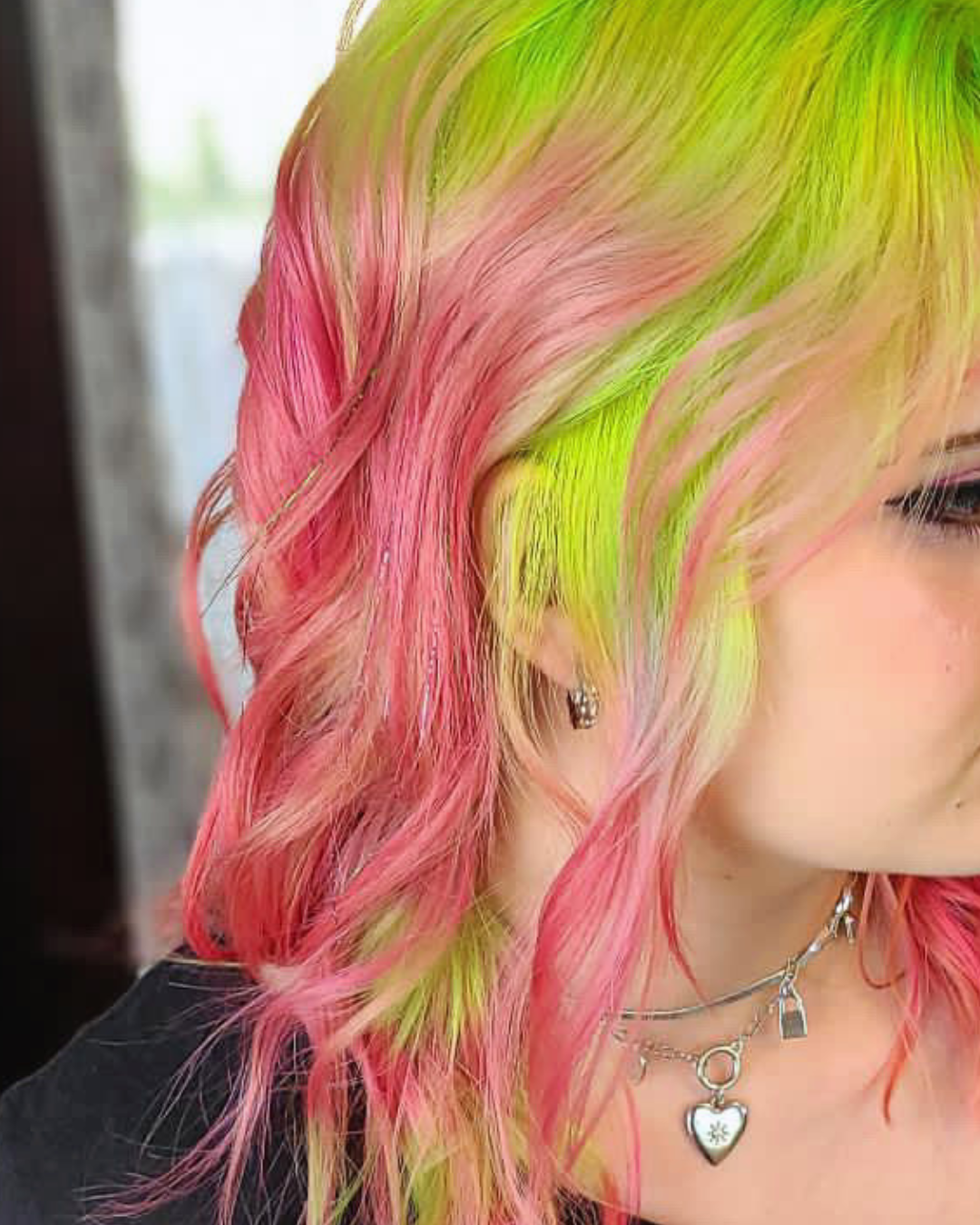 Close-up of a woman's multi-colored hair with pink, yellow, and green shades, wearing earrings and layered necklaces.