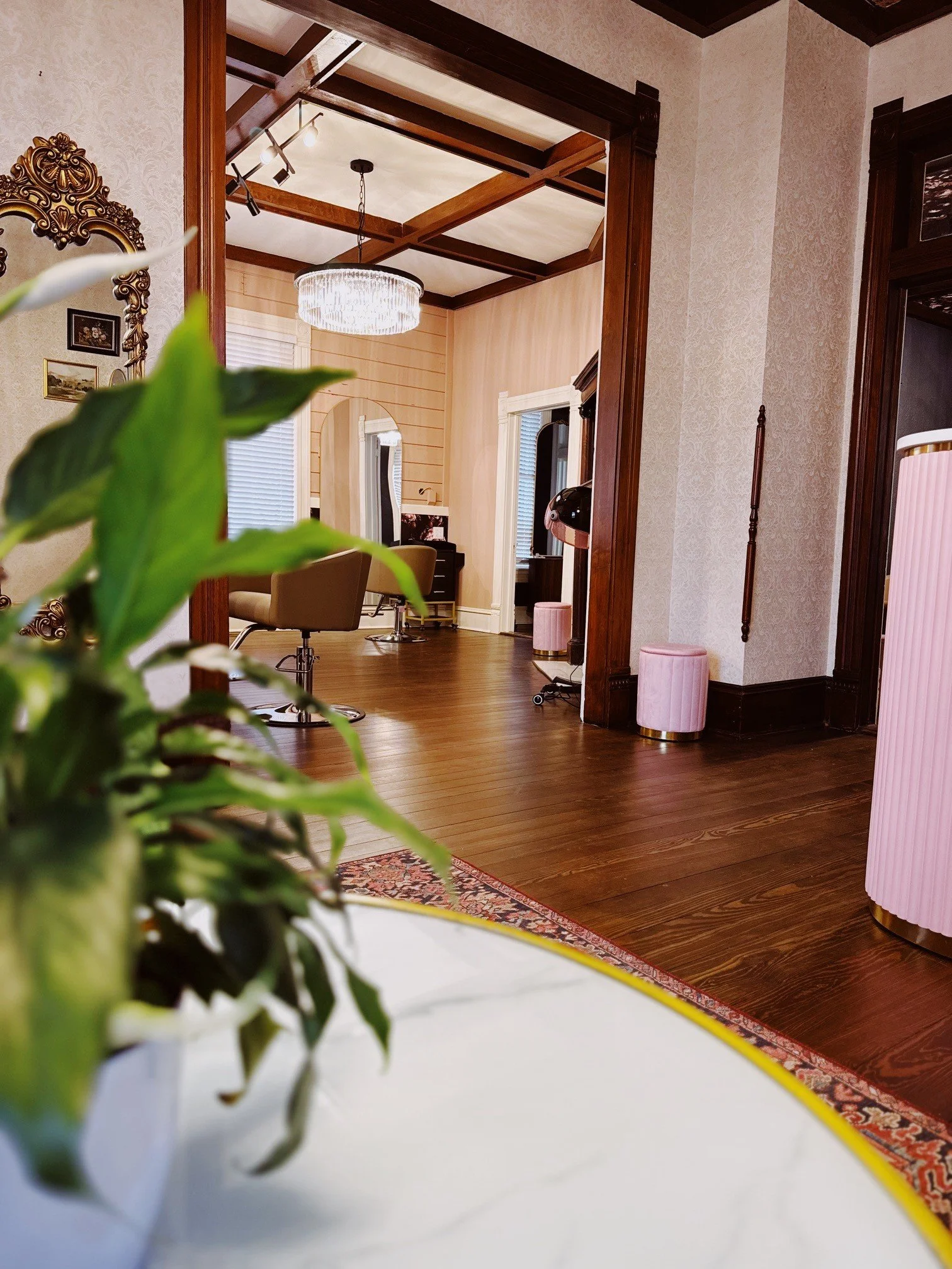 Interior view of a room with dark wooden floor, decorative molding, a pink ottoman, a pink cylindrical object, and a chandelier hanging from the ceiling.