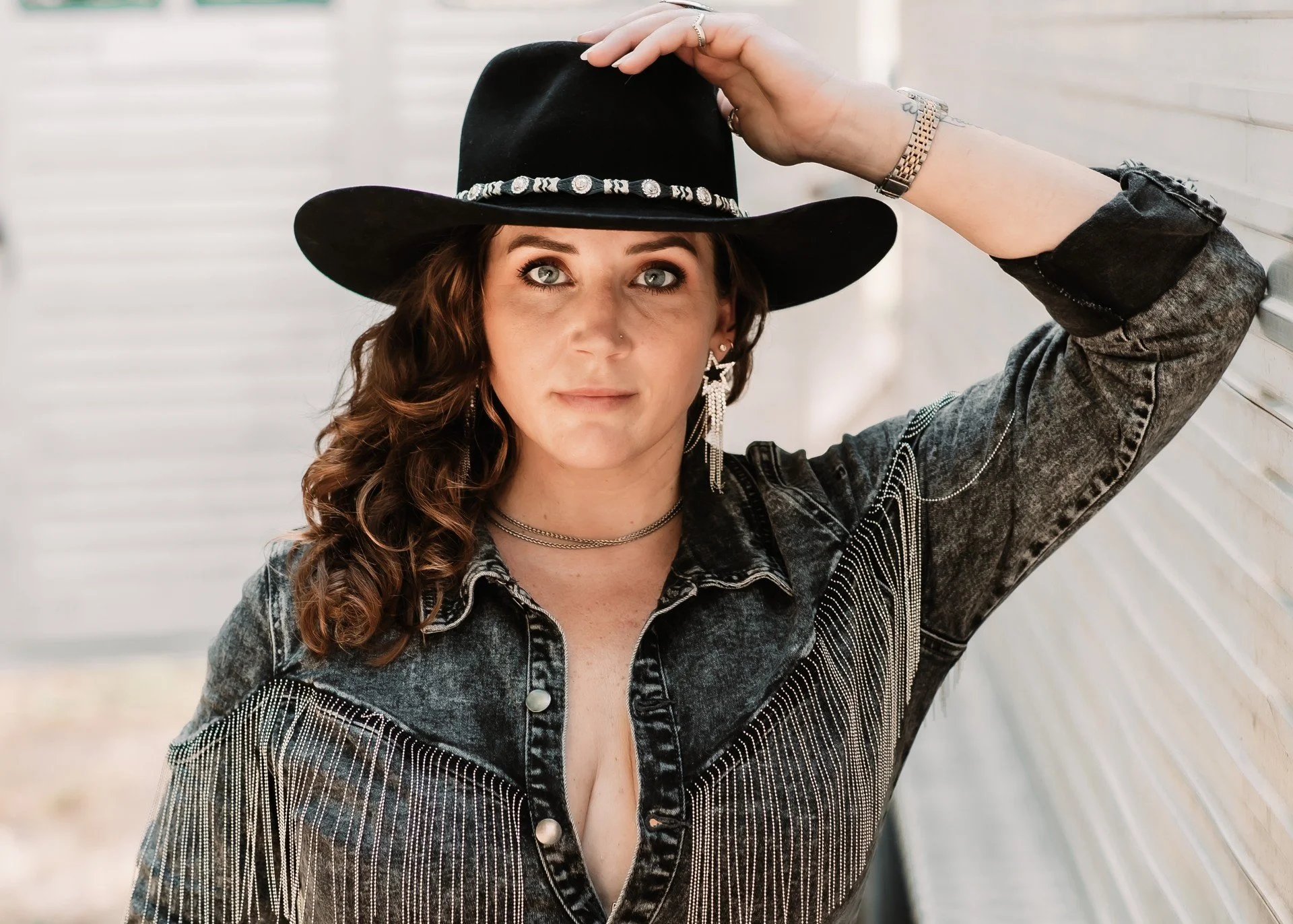 A woman with wavy brown hair wearing a black wide-brim hat, denim jacket with embellishments, and silver jewelry, standing next to a white wooden wall.