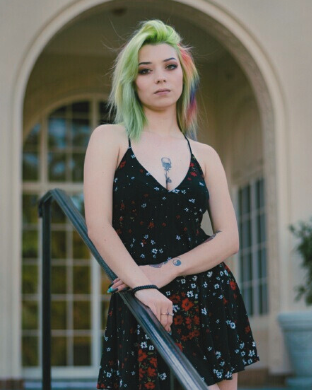 Young woman with green, yellow, and pink hair standing outdoors in front of a building with large arches. She is wearing a black dress with a floral pattern and has visible tattoos on her chest, neck, and arm.