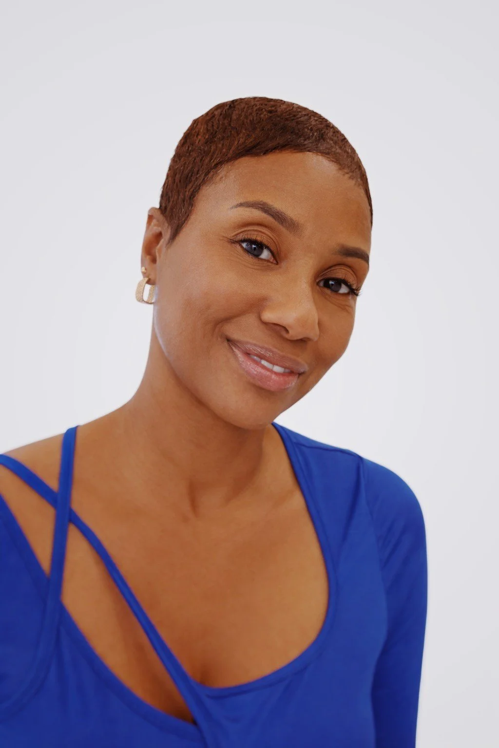 A woman smiling with short reddish-brown hair, wearing hoop earrings and a blue top with thin straps, against a plain white background.