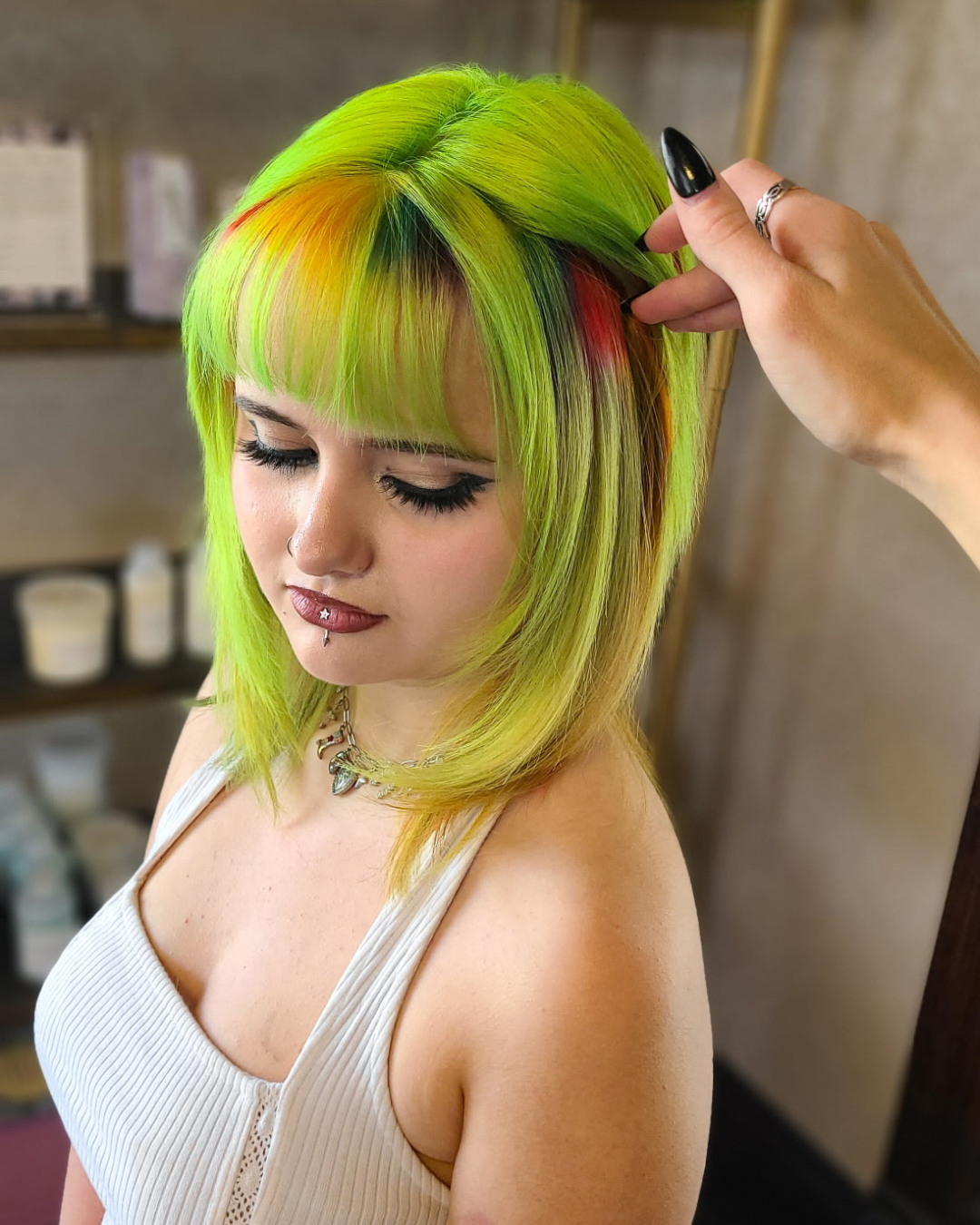 Young woman with vibrant green, yellow, and red hair, wearing a white sleeveless top, inside a salon or studio, having her hair styled.