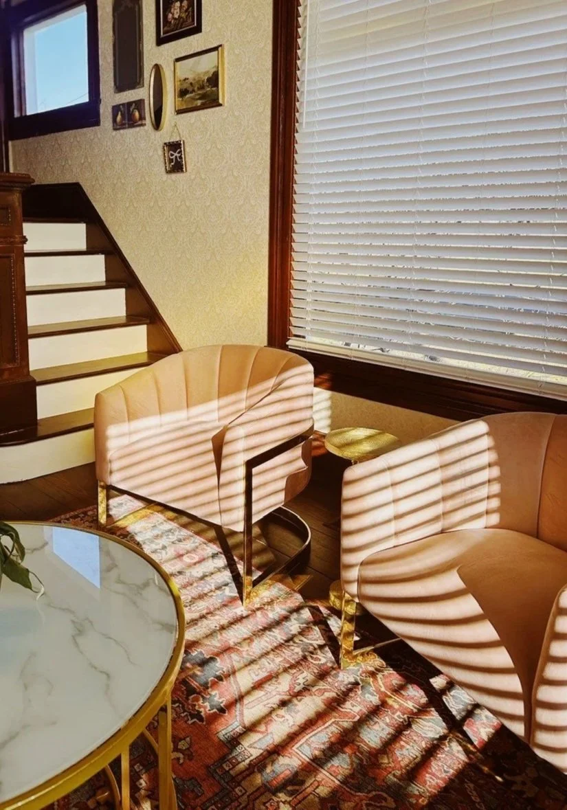 Sunlight streams through window blinds, casting striped shadows on two peach-colored armchairs, a small gold side table, a marble-topped dining table, a patterned rug, and a wall with framed pictures.