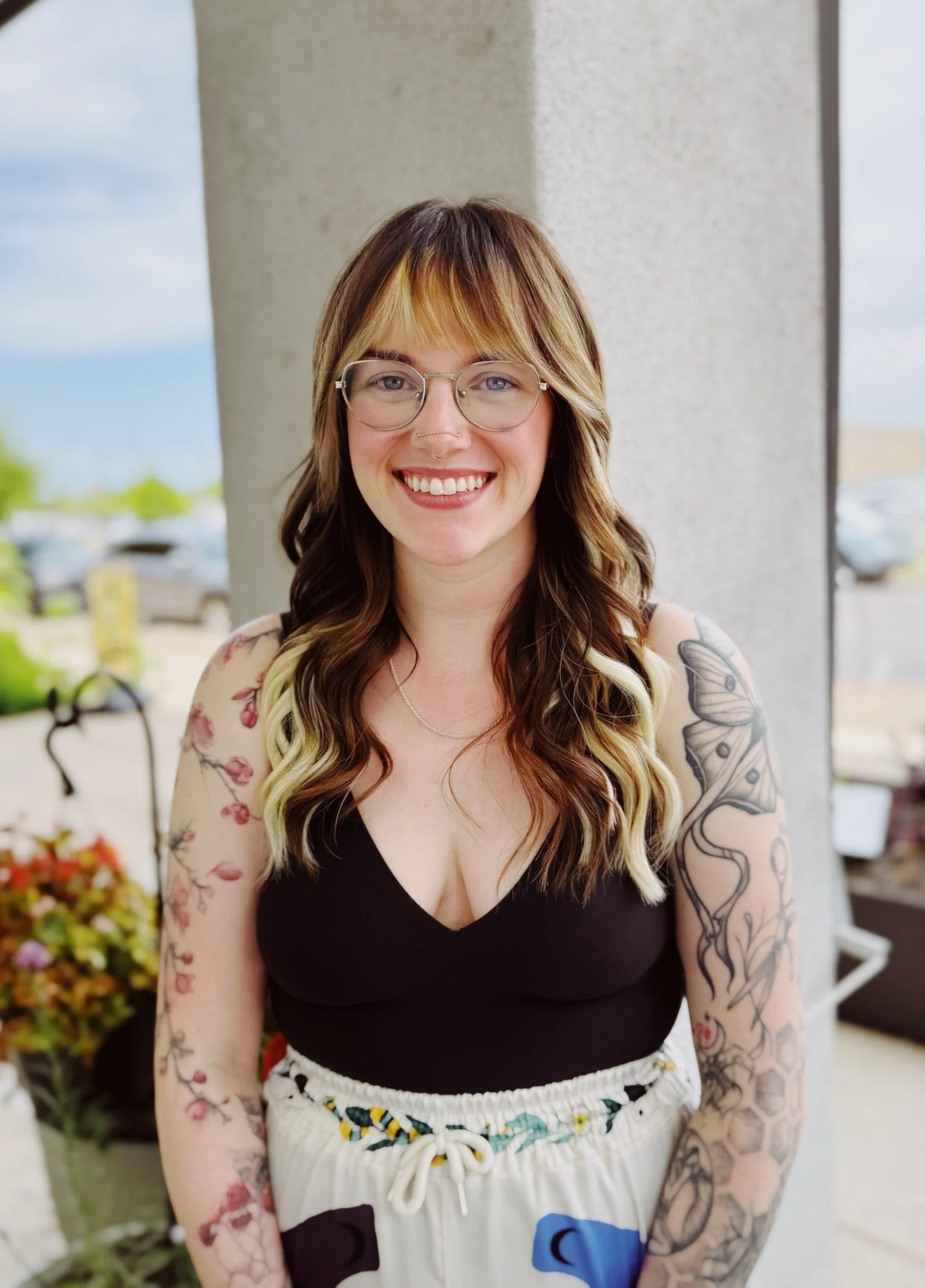 A smiling woman with long wavy hair, glasses, and tattoos on her arms, standing outdoors near a concrete pillar with a parking lot and blue sky in the background.