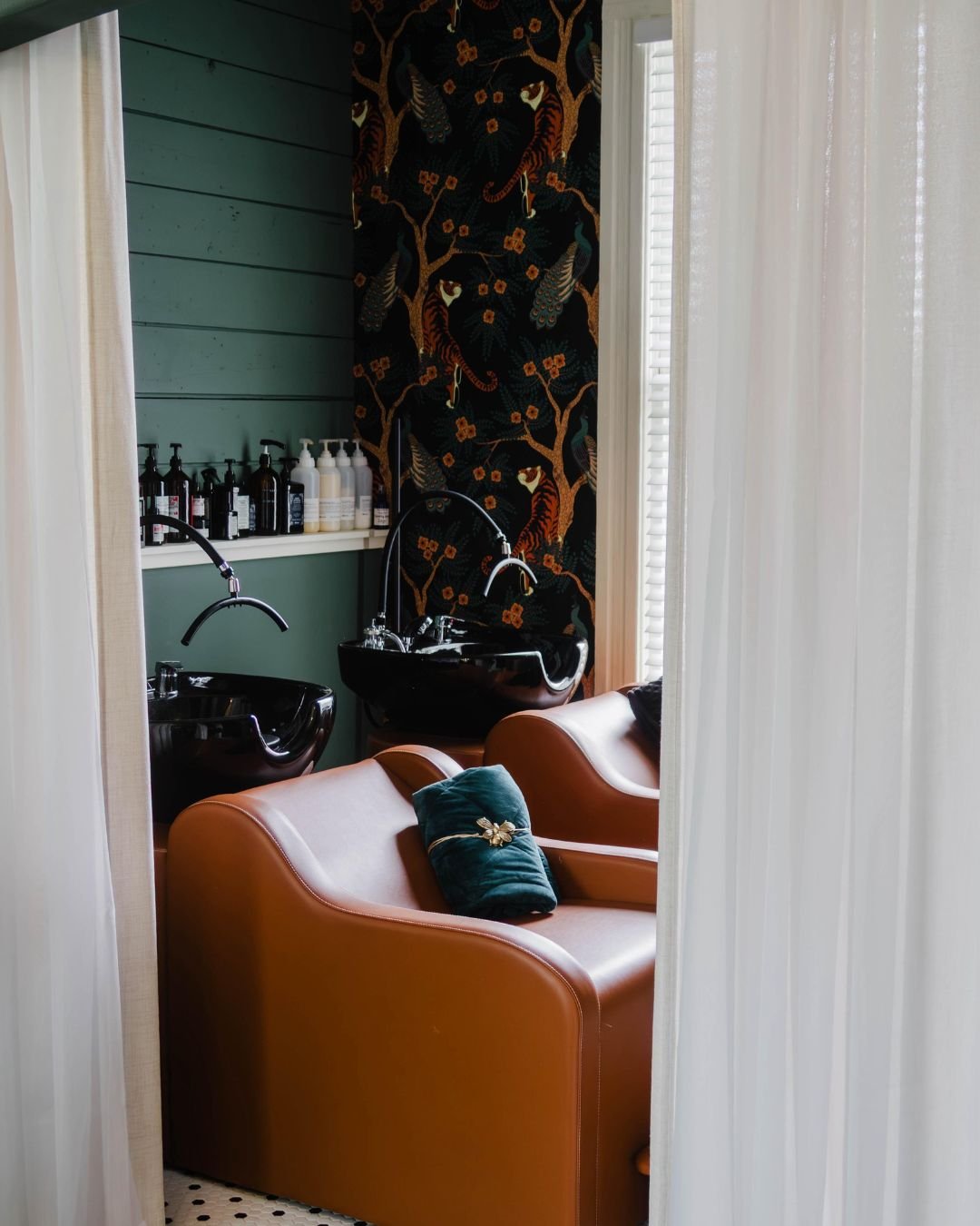 Bathroom with dark wallpaper featuring floral patterns, stained glass windows with colorful abstract designs, a mirror reflecting the window, a marble-topped vanity with a silver frame, a faucet, a bottle of soap, a toilet, and a roll of toilet paper.