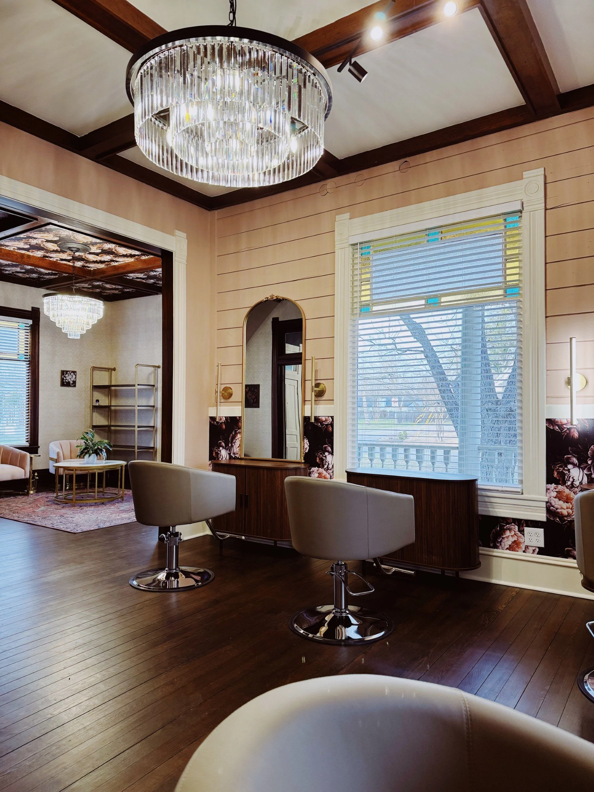 Interior of a hair salon with styling chairs, mirror, large window, and chandelier lighting.